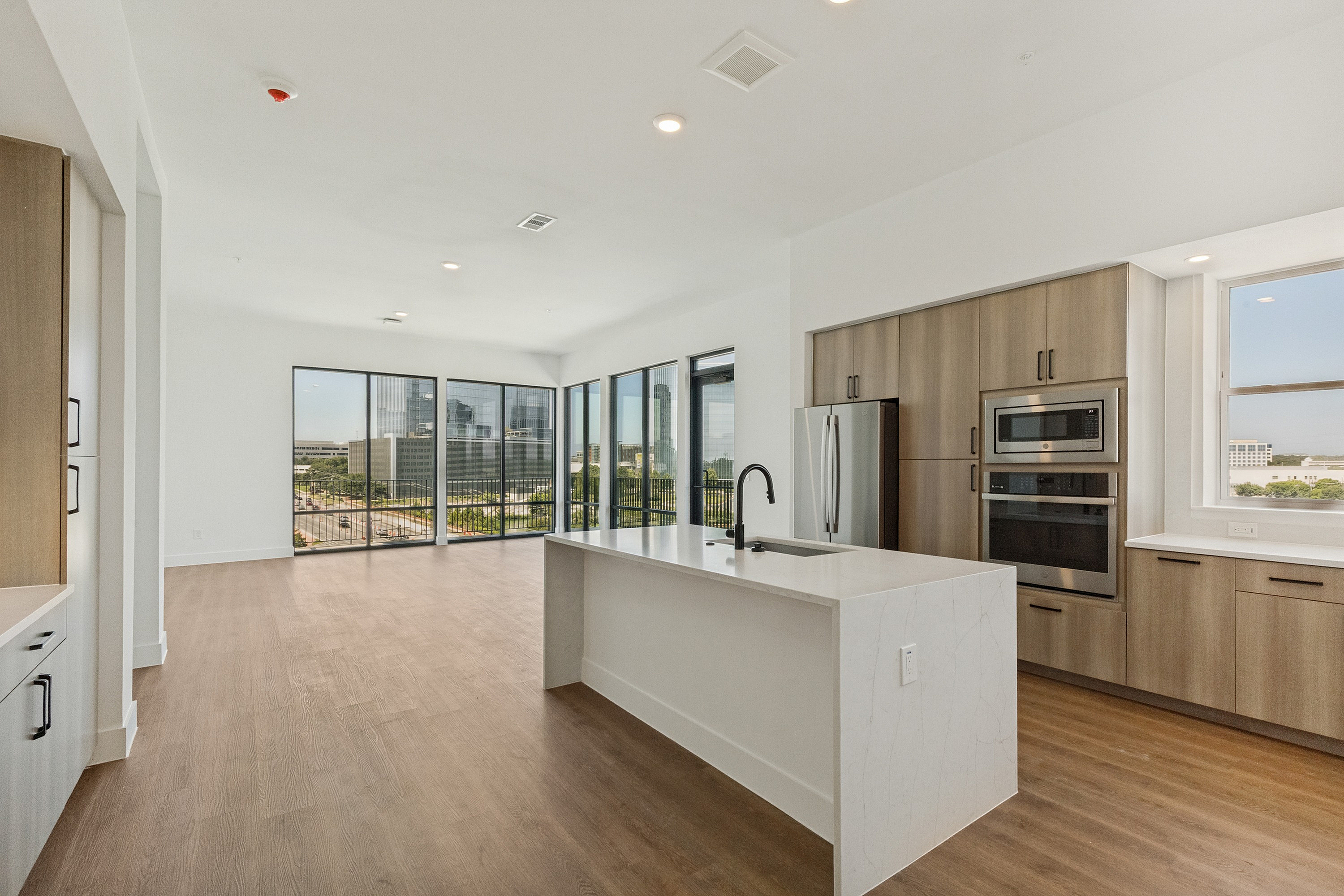 Bright open-concept apartment with sleek kitchen island, ample wood cabinetry, and floor-to-ceiling windows offering stunning city views.