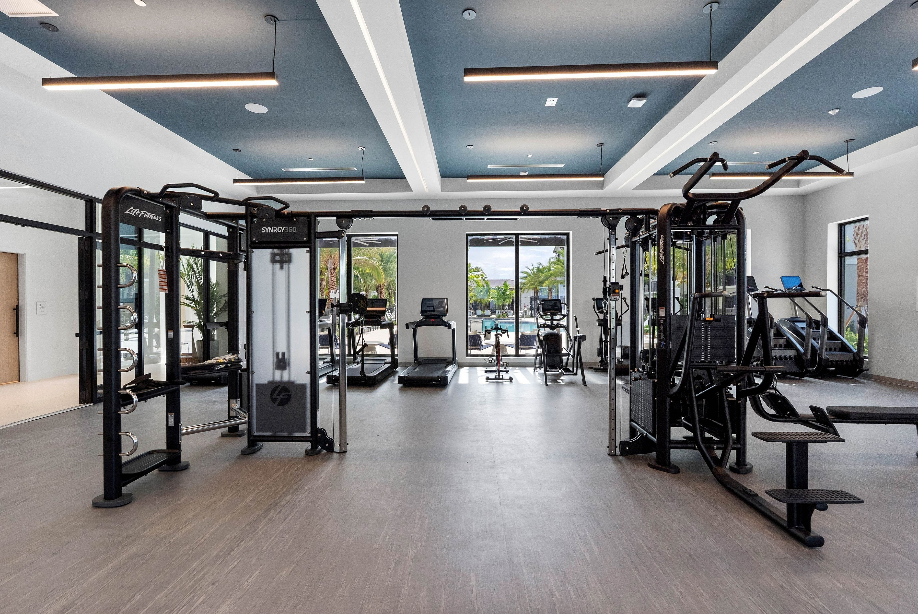 Modern fitness center with state-of-the-art equipment, large windows offering natural light, and views of a tranquil outdoor pool area.