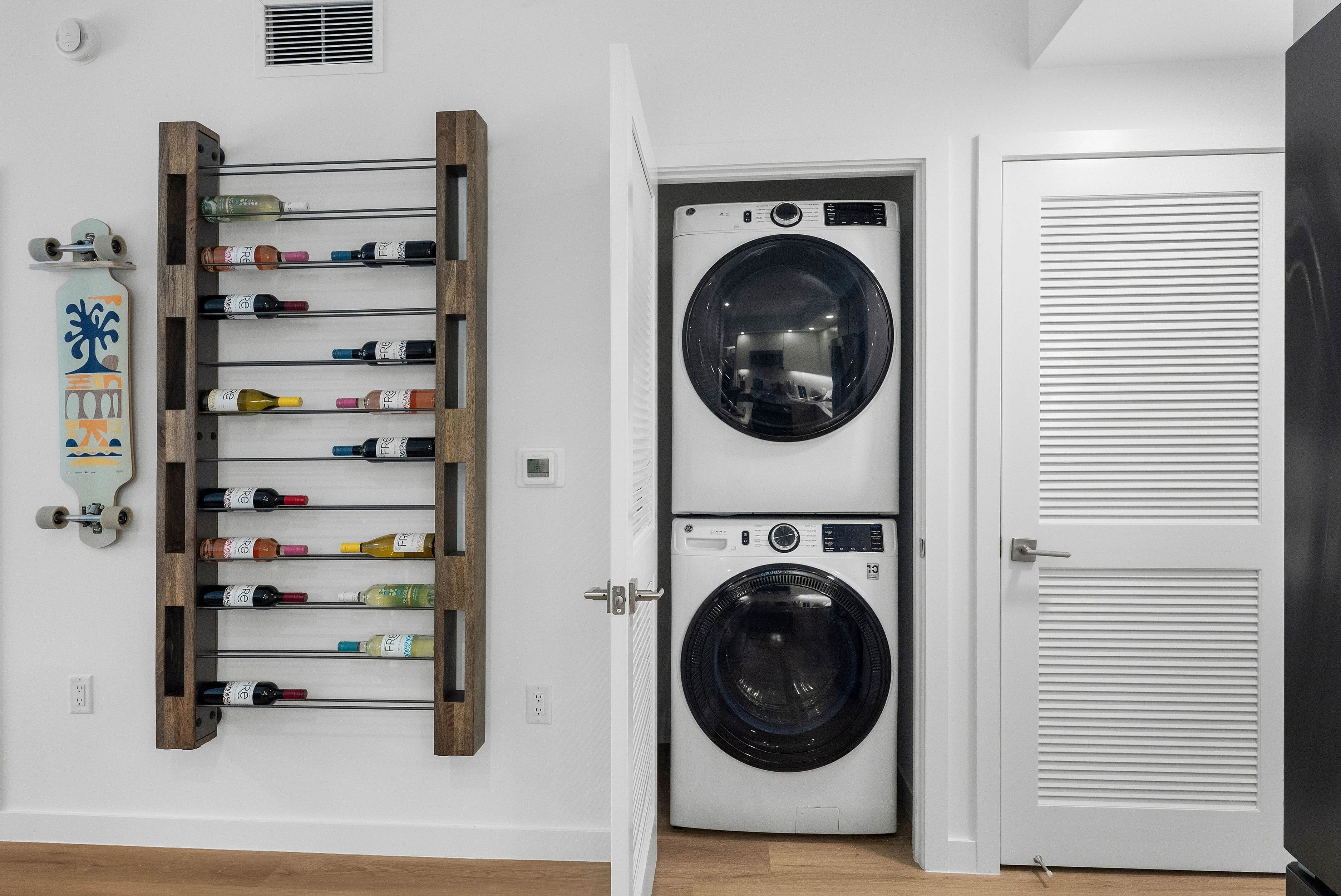 Contemporary apartment interior featuring a sleek stacked washer and dryer, a stylish wooden wine rack, and vibrant skateboard art.