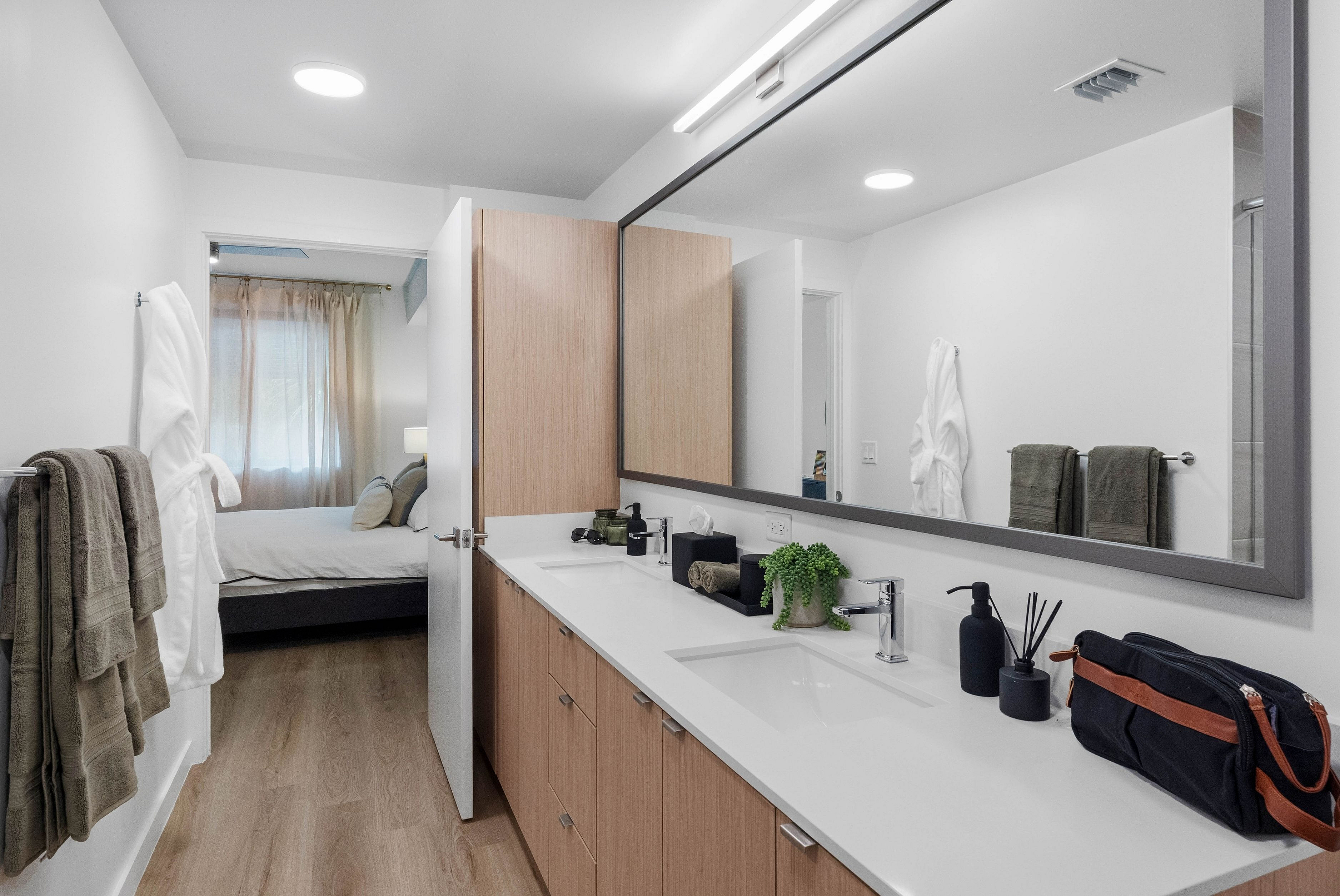 Modern bathroom with sleek double vanity, chic wooden cabinetry, and view into a cozy, sunlit bedroom.