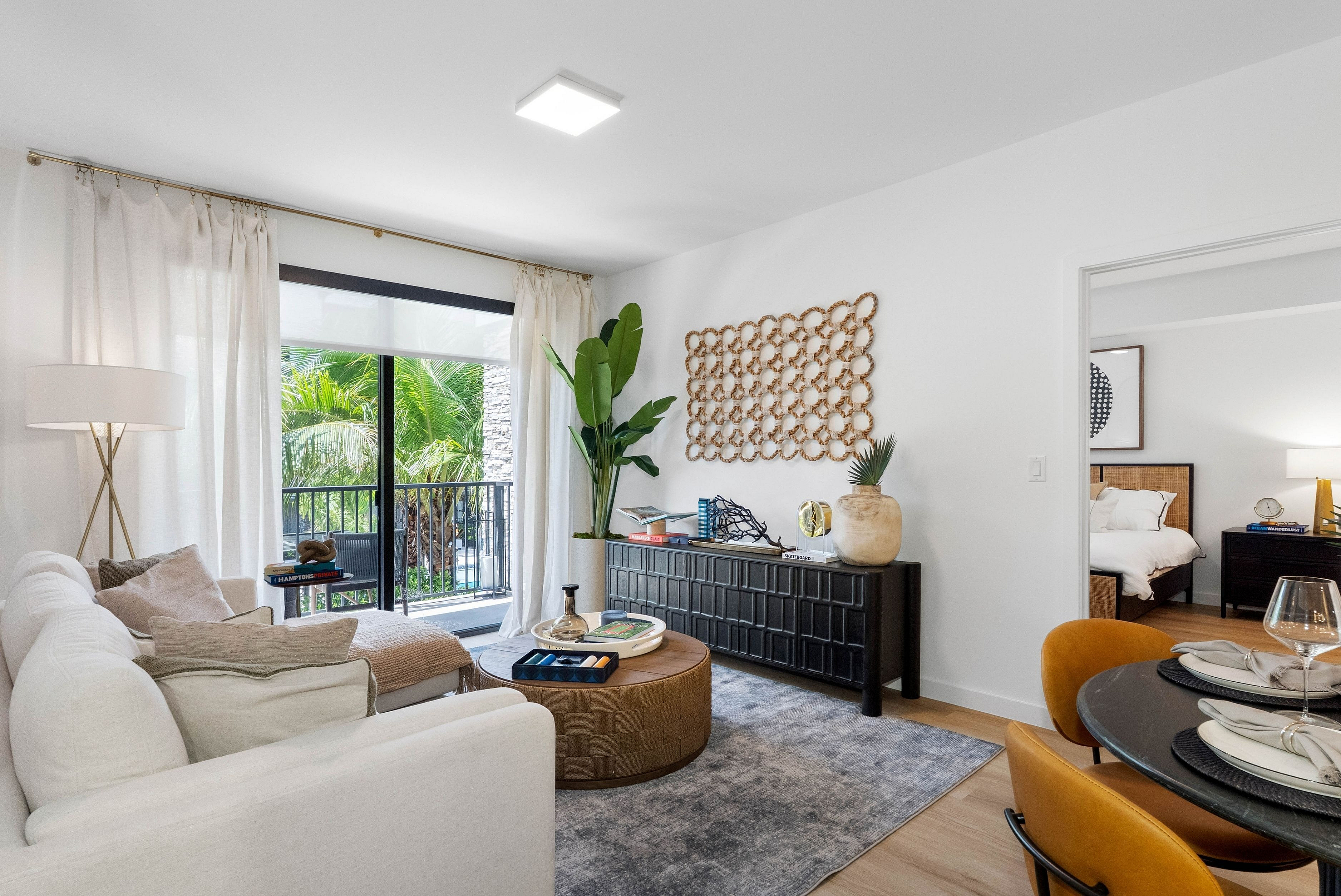 Modern living room with elegant décor, large windows allowing natural light, cozy seating, decorative plants, and a glimpse into a stylish bedroom.