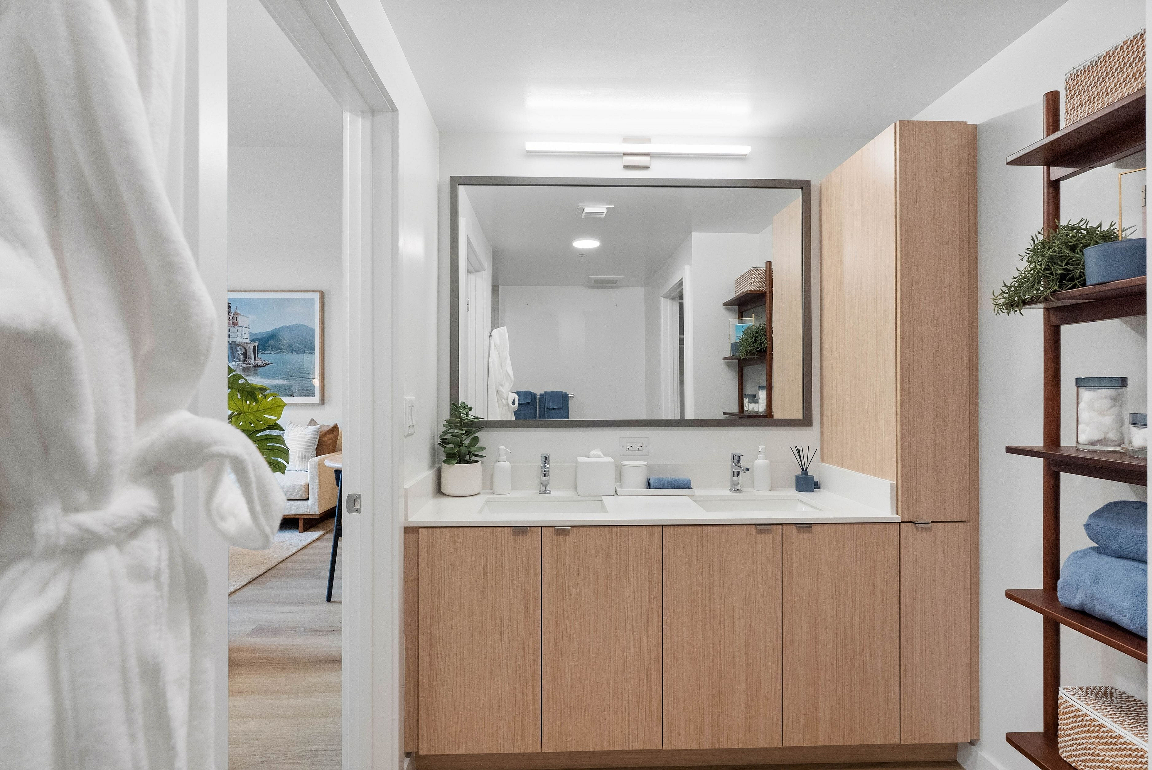 Modern bathroom with sleek wooden cabinetry, large mirror, and plush decor leading to a cozy living area.