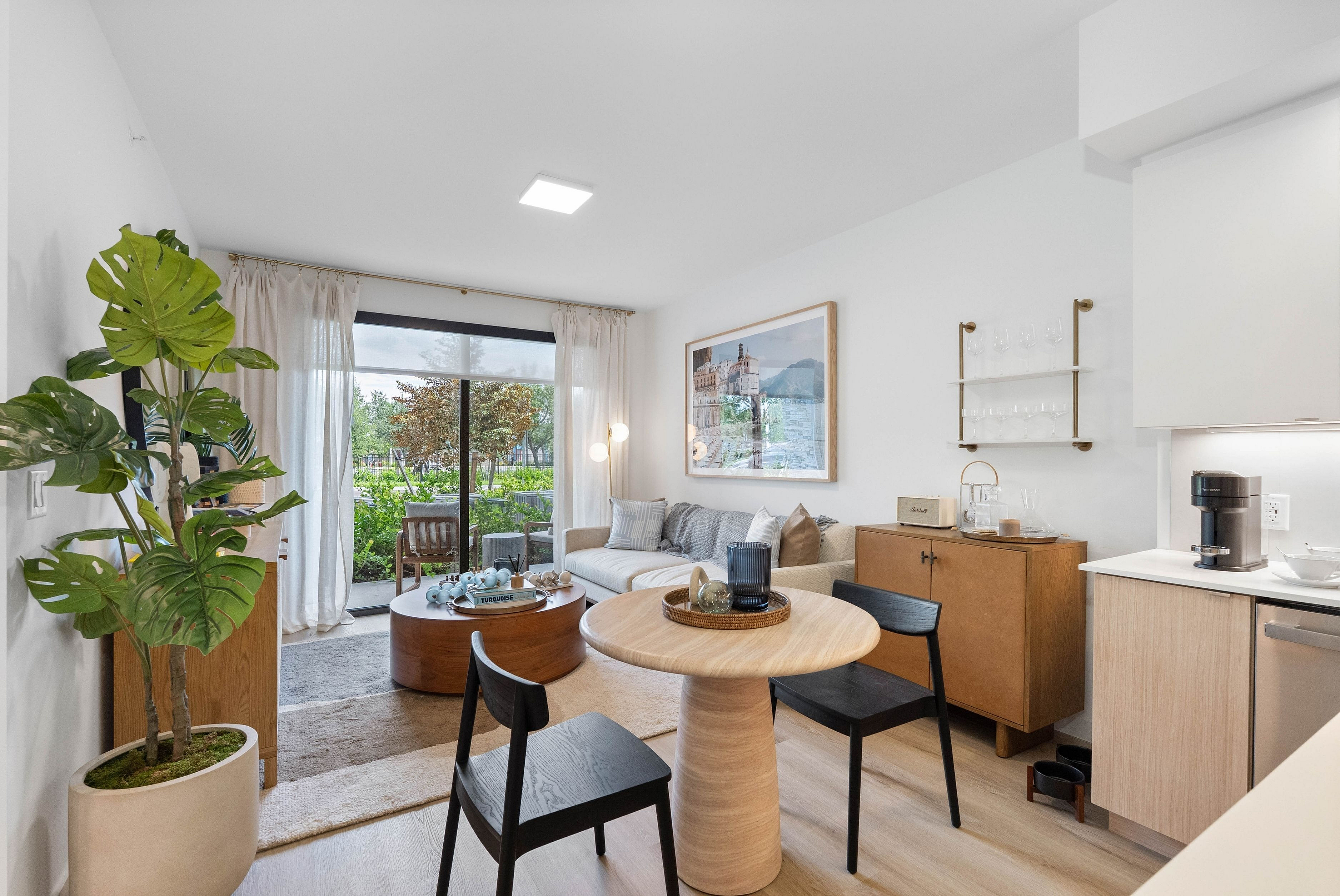 Bright and modern open-concept living and dining area with stylish decor, featuring a cozy sofa, elegant artwork, and lush greenery with a view through large glass doors.