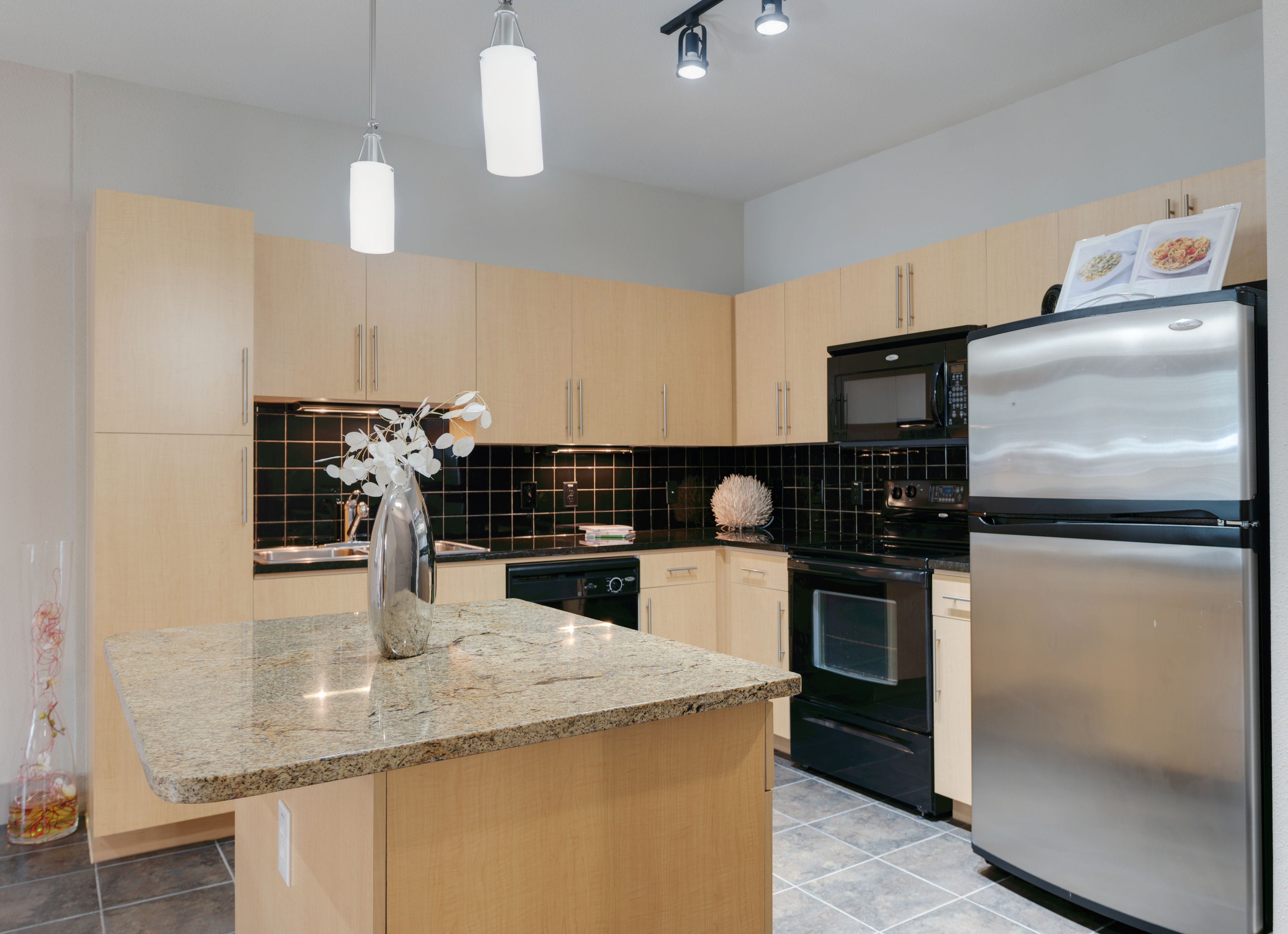 Kitchen with granite island, black tile backsplash, light wood cabinets, and stainless steel refrigerator