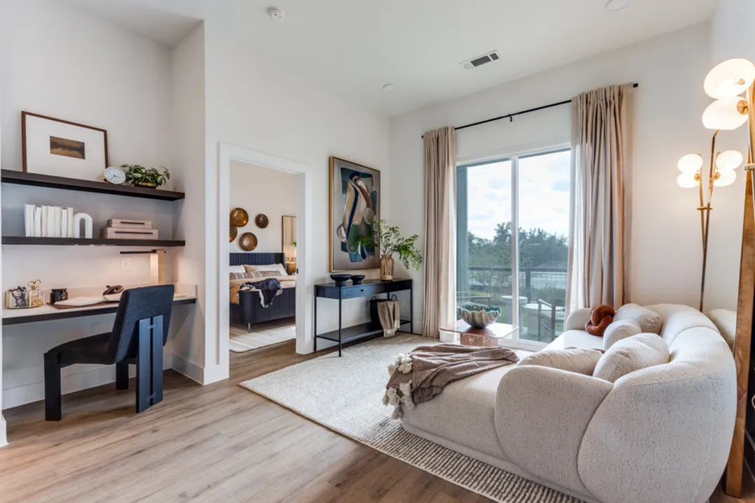 Elegant living space with cozy seating, a dedicated workspace, and large windows inviting natural light; bedroom access through a stylish doorway.