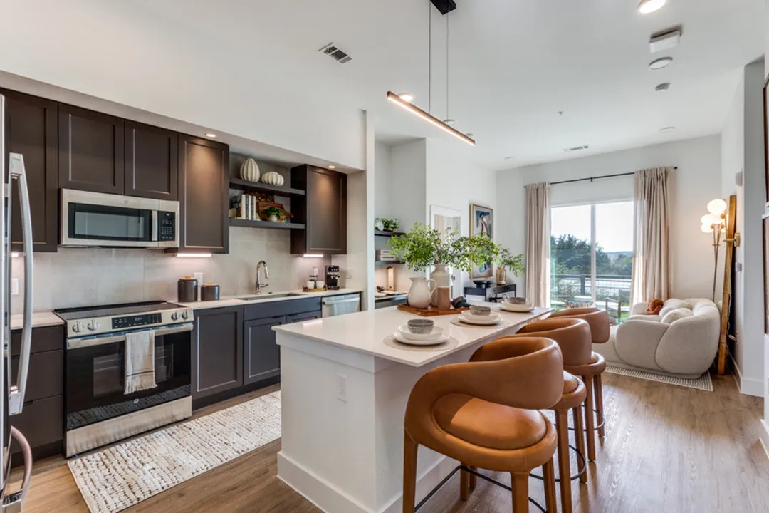 Chic corner kitchen with dark cabinets, modern appliances, and cozy bar seating opening to a bright living area with lush garden views.