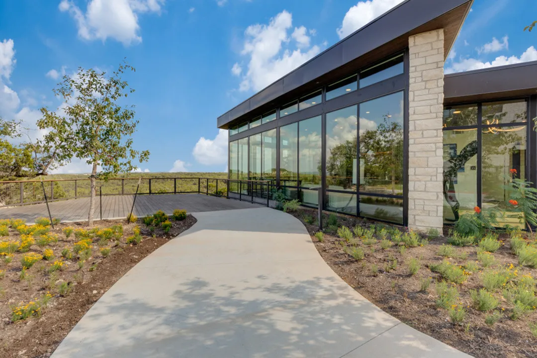 Modern building with expansive glass windows, surrounded by lush landscaping and a serene pathway under a bright blue sky.
