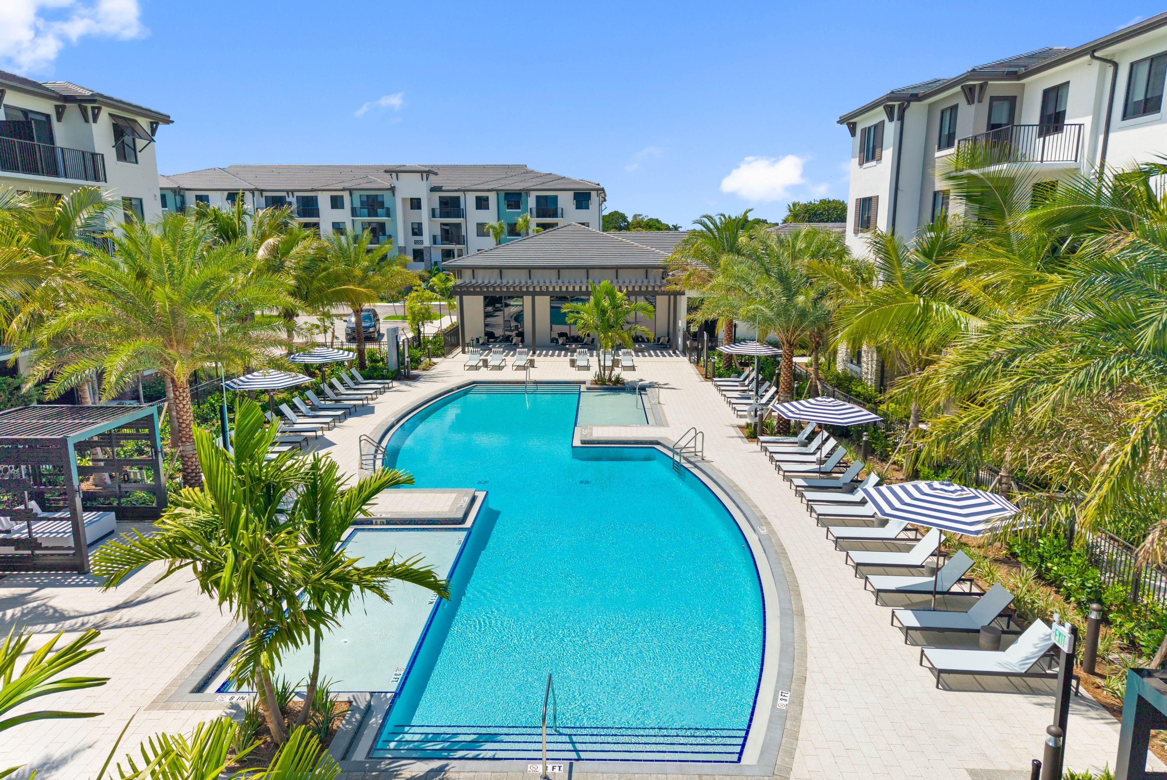 Luxurious apartment complex featuring a stunning blue pool surrounded by lush palm trees and elegant lounge chairs.