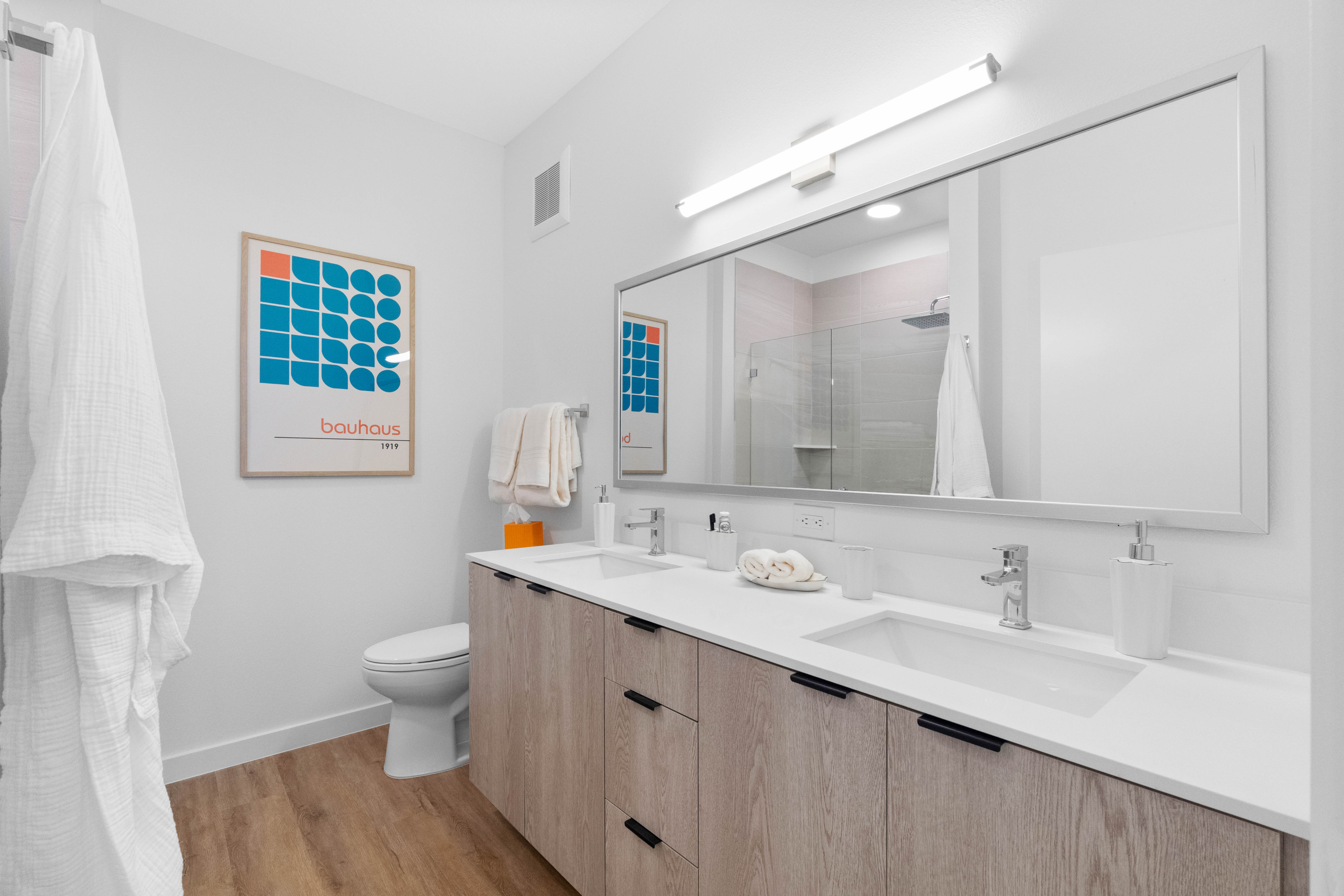 Modern bathroom with elegant double vanity, stylish wall art, and sleek glass shower, featuring natural wood flooring.