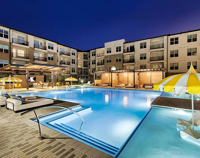 Beautifully lit apartment complex with a luxurious pool, lounge chairs, and yellow striped umbrellas at night.