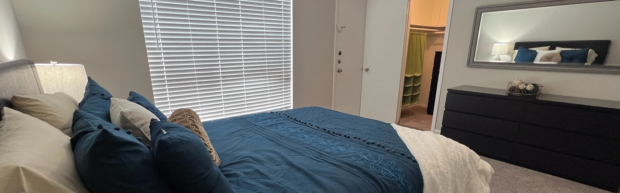 A modern bedroom featuring a comfortable blue bed, stylish dresser, and large window with blinds.