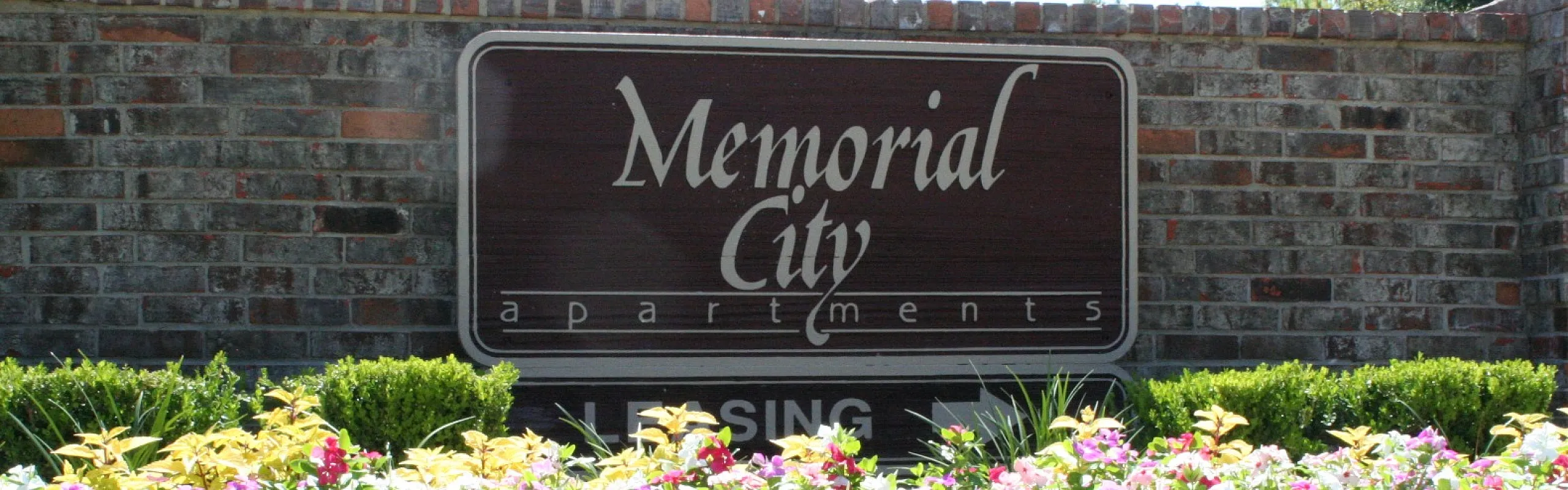 Entrance to Memorial City Apartments with vibrant flower beds and brick wall signage.