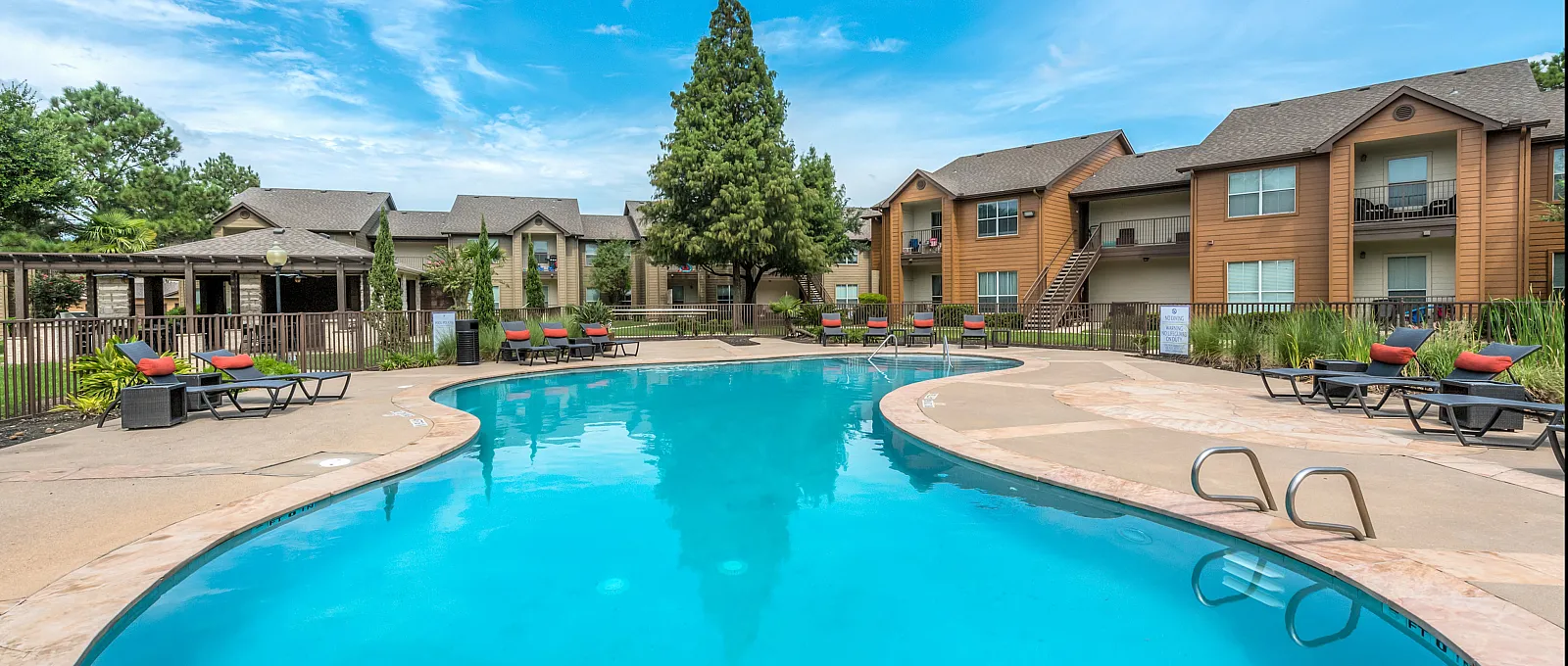 Inviting apartment complex showcasing a sparkling blue pool surrounded by lush greenery and elegant lounge chairs under a bright blue sky.