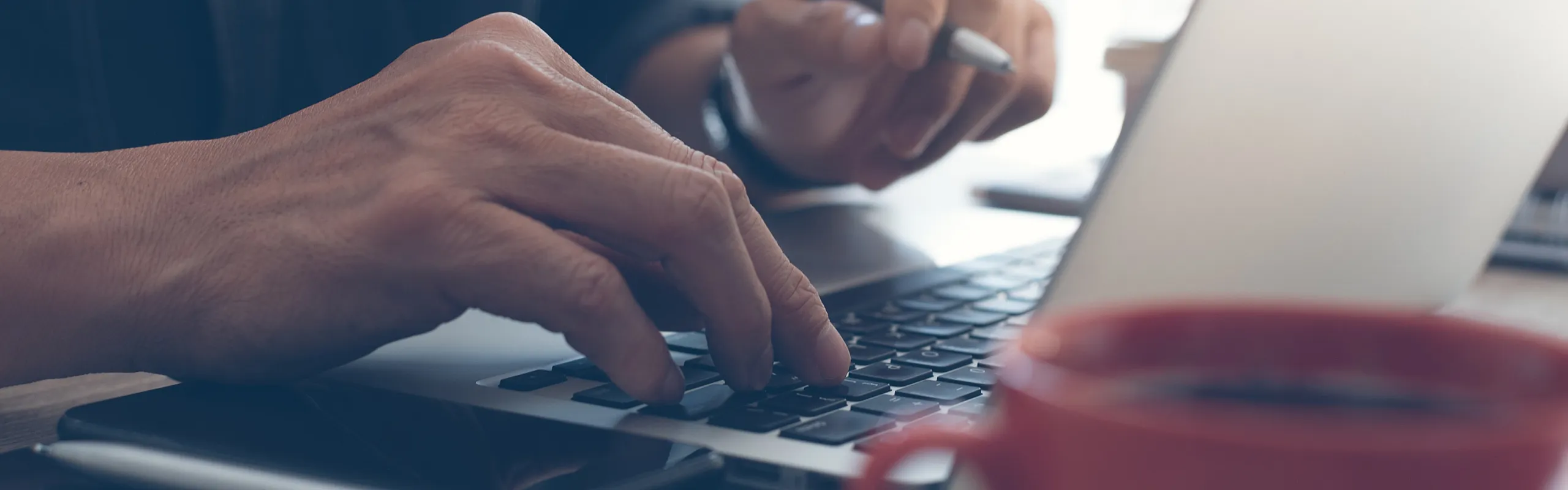 Focused workspace featuring a person typing on a laptop with a pen, tablet, and coffee cup nearby, conveying productivity and efficiency.