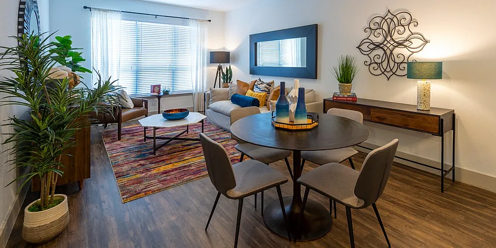 Inviting living and dining area with modern decor, featuring a cozy sofa, vibrant rug, and elegant dining set, bathed in natural light from large windows.