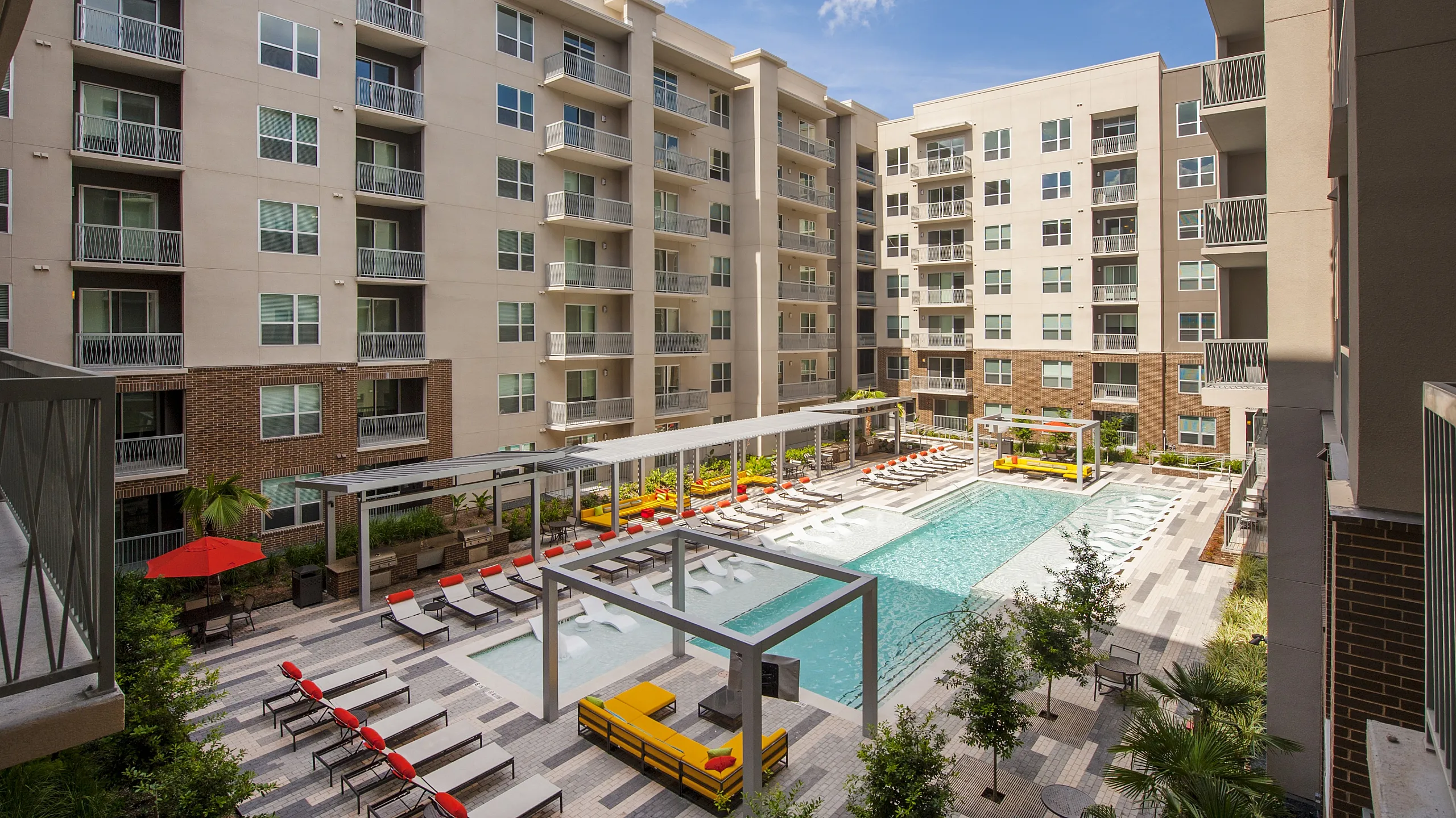 Modern apartment complex featuring luxurious swimming pool, stylish lounge areas, and sunlit balconies surrounded by lush greenery.