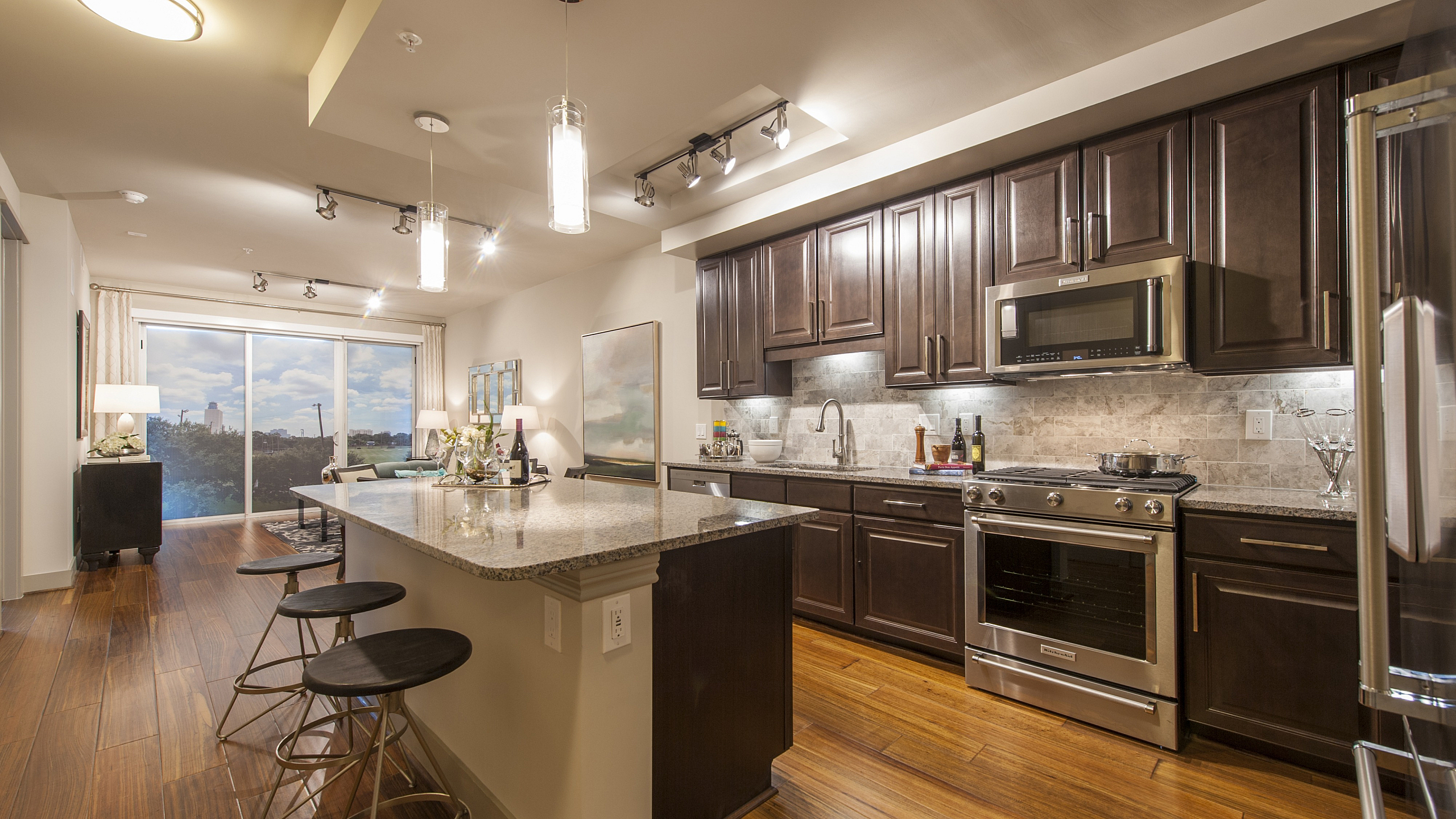 Modern kitchen with sleek dark wood cabinetry, stainless steel appliances, and a granite island with stylish bar stools, overlooking a bright living area with floor-to-ceiling windows.