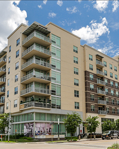 Modern seven-story apartment complex with spacious balconies, surrounded by lush greenery, set against a vibrant blue sky.