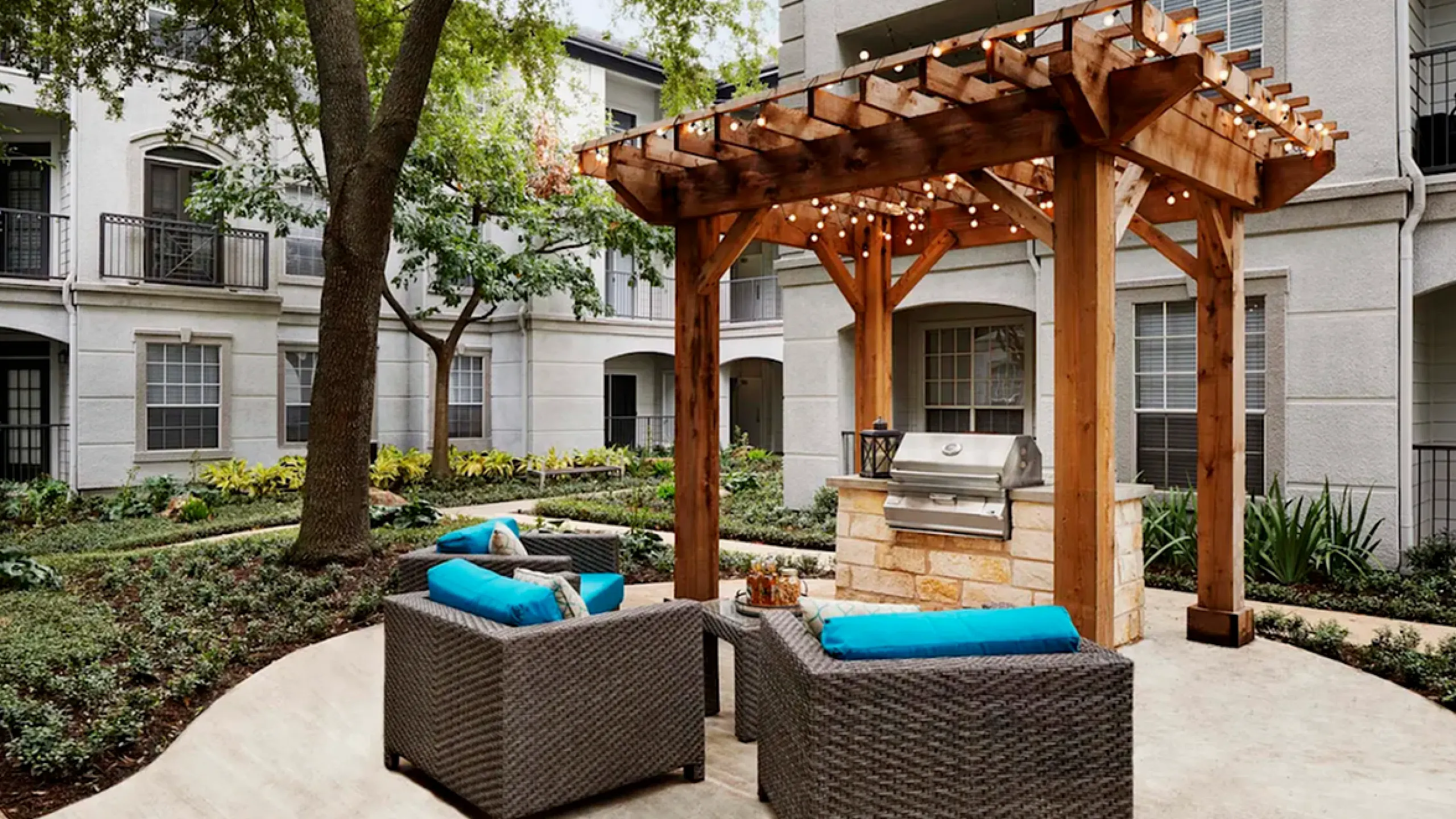 Charming apartment courtyard featuring cozy wicker seating with vibrant blue cushions, modern grill under a stylish pergola, surrounded by lush greenery.