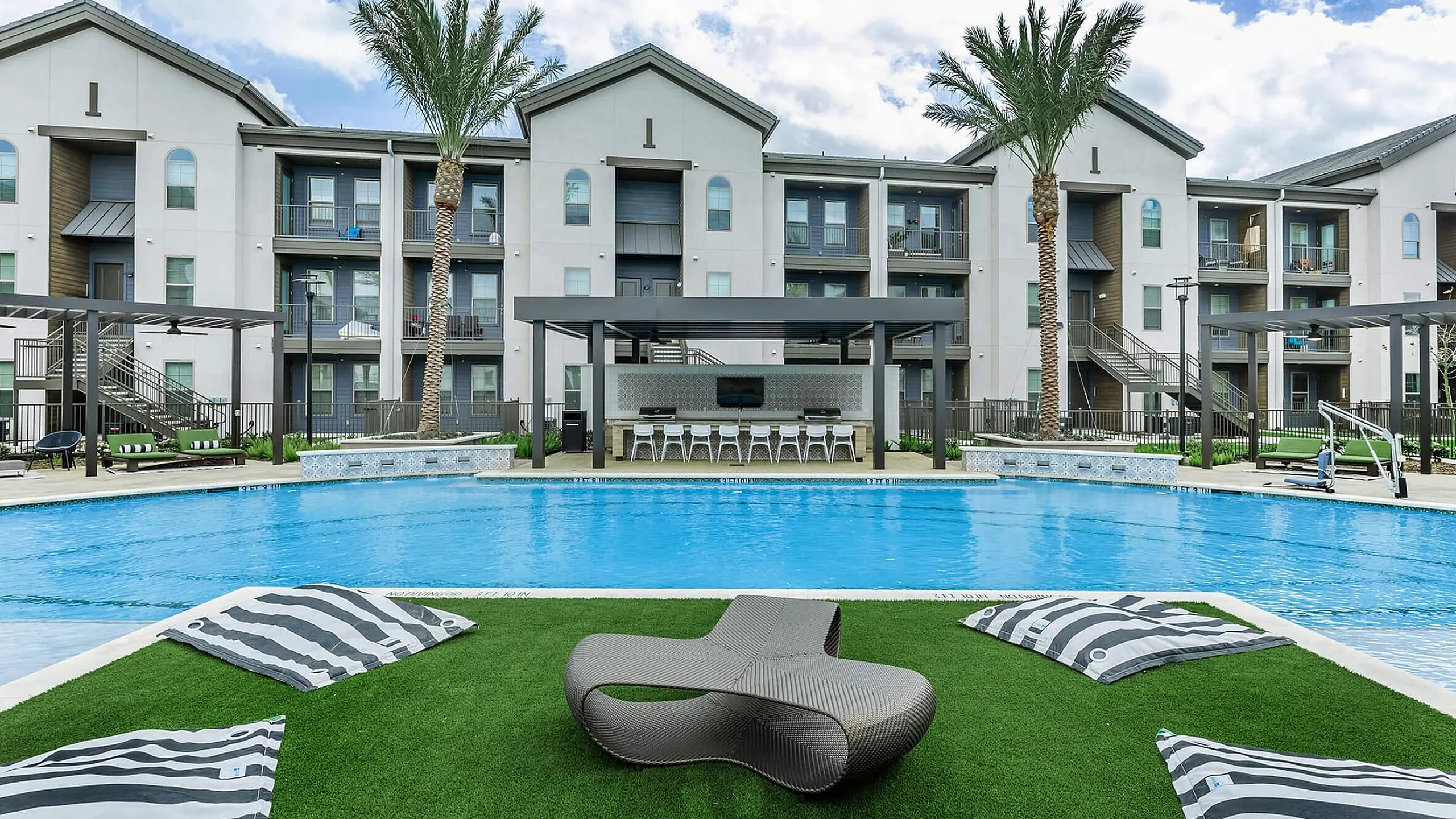 Luxurious apartment complex showcasing a sparkling pool, stylish lounge areas, and modern architecture under a vibrant blue sky.
