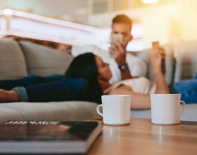 Cozy living room with a soft couch and a relaxing couple, featuring warm natural light and two stylish coffee mugs on a wooden table.