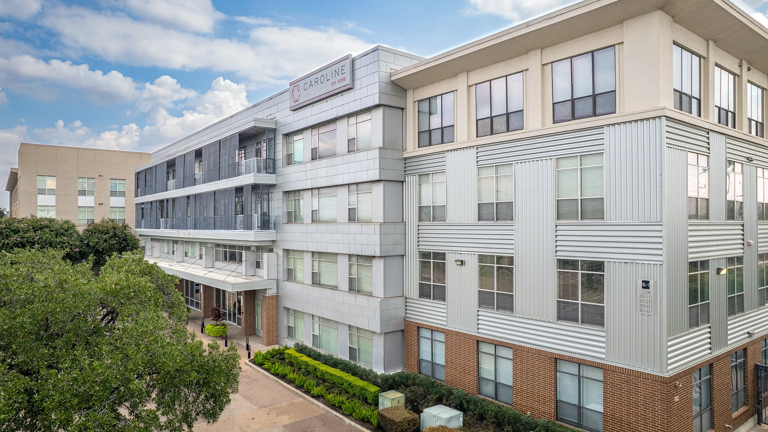 Modern apartment complex with a sleek architectural design featuring large windows, lush greenery, and a welcoming entrance under a blue sky.