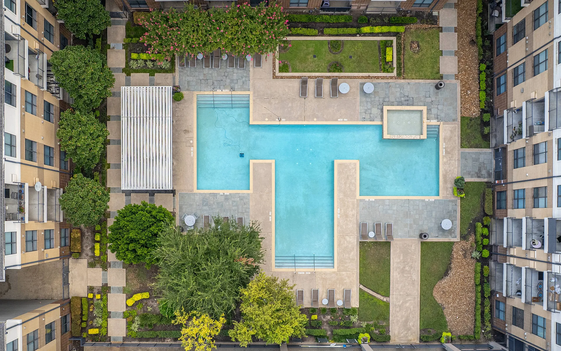 Aerial view of a luxurious apartment complex featuring a large T-shaped pool surrounded by lush greenery, modern lounge chairs, and pristine landscaping.