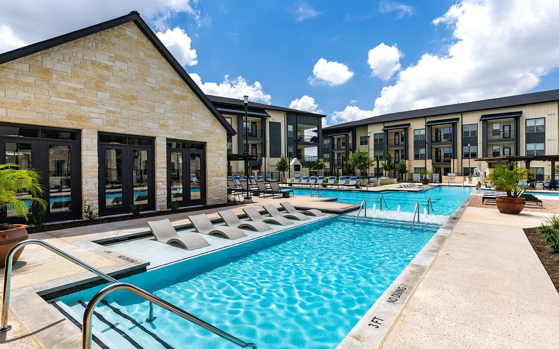 Luxurious apartment complex with a stunning outdoor pool area, featuring modern lounge chairs and a vibrant, clear blue sky.