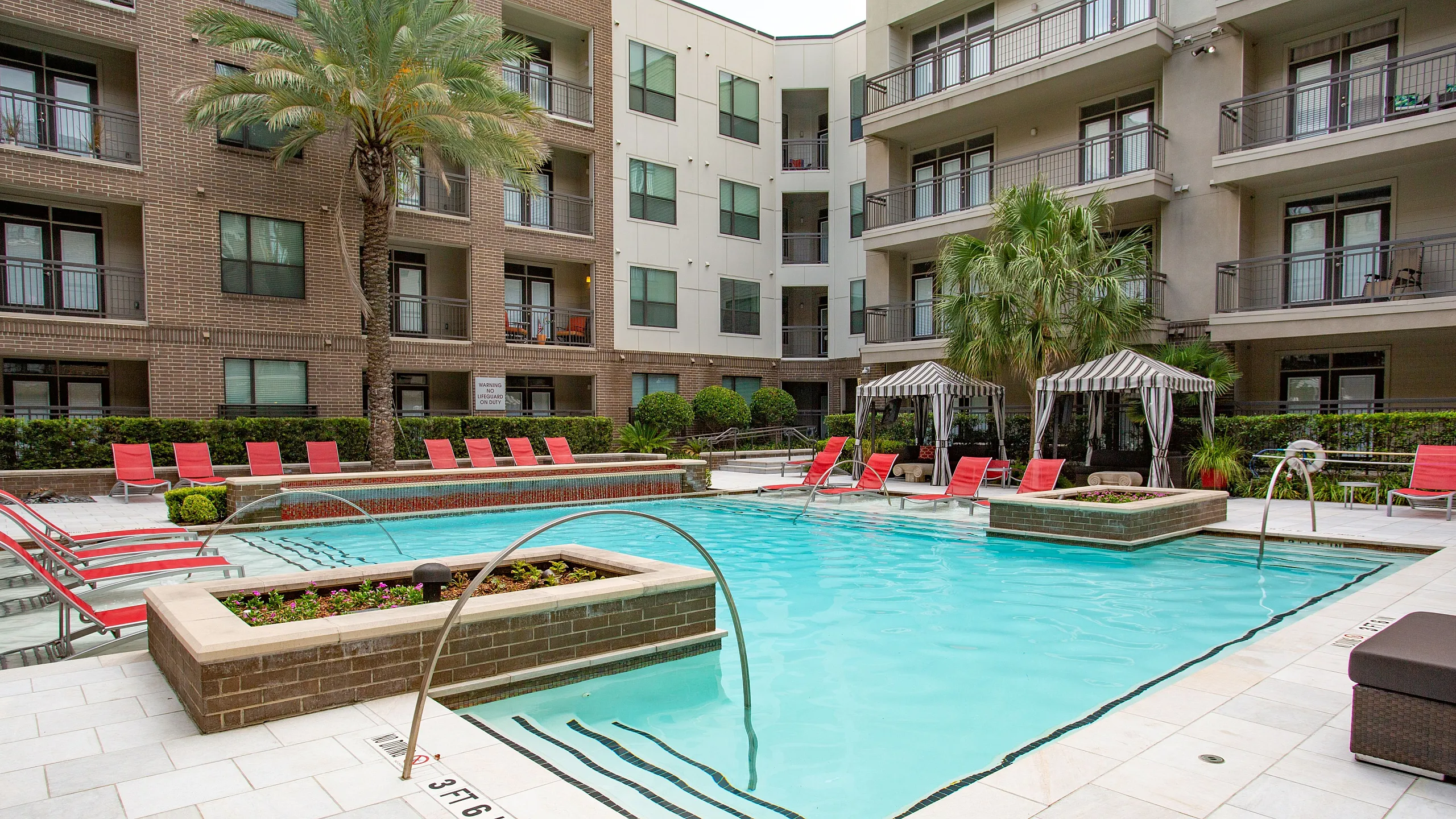 Luxurious apartment complex courtyard featuring a sparkling swimming pool with vibrant red lounge chairs, shaded private cabanas, and lush palm trees.