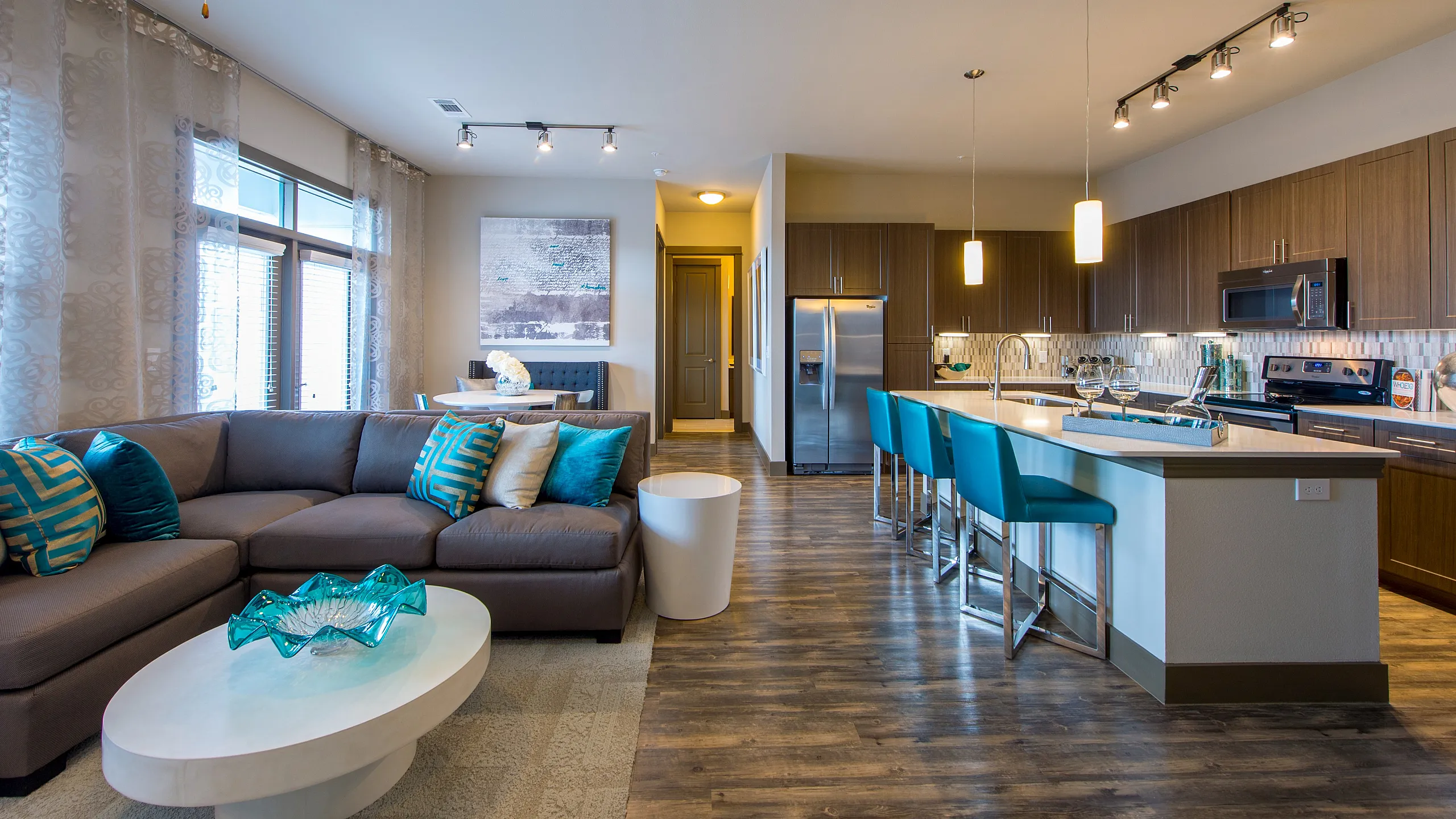 Modern open-concept living area featuring a stylish kitchen with teal accents, cozy sectional sofa, and well-lit dining space.