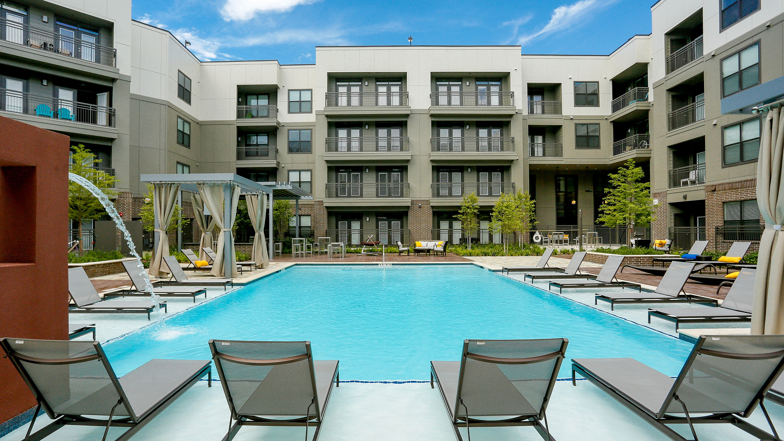Luxurious apartment complex with a sparkling pool, elegant cabanas, and stylish lounge chairs under a vibrant blue sky.