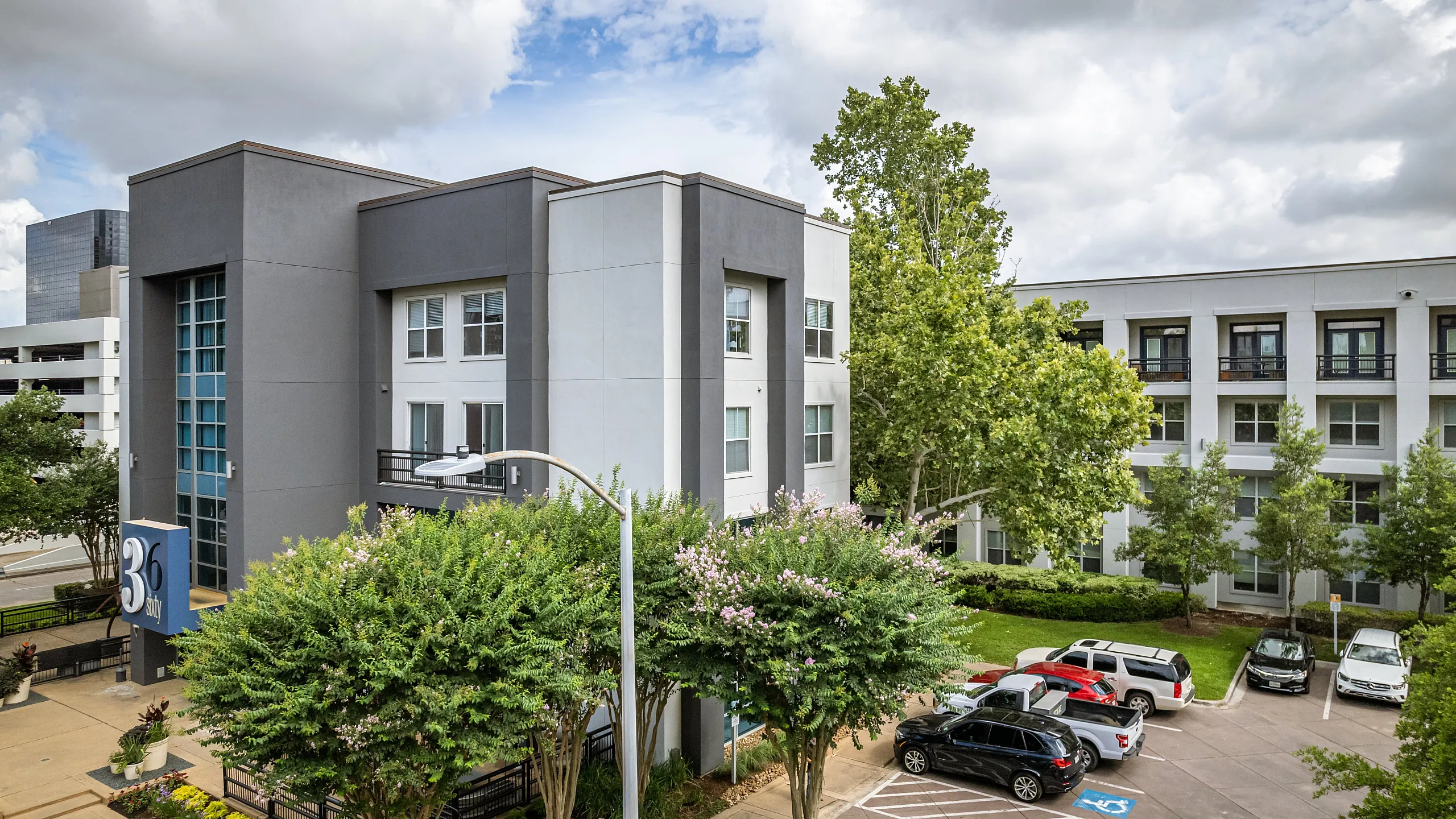 Modern apartment complex with sleek architectural design, lush landscaping, and ample parking, set against a backdrop of vibrant greenery and a partly cloudy sky.