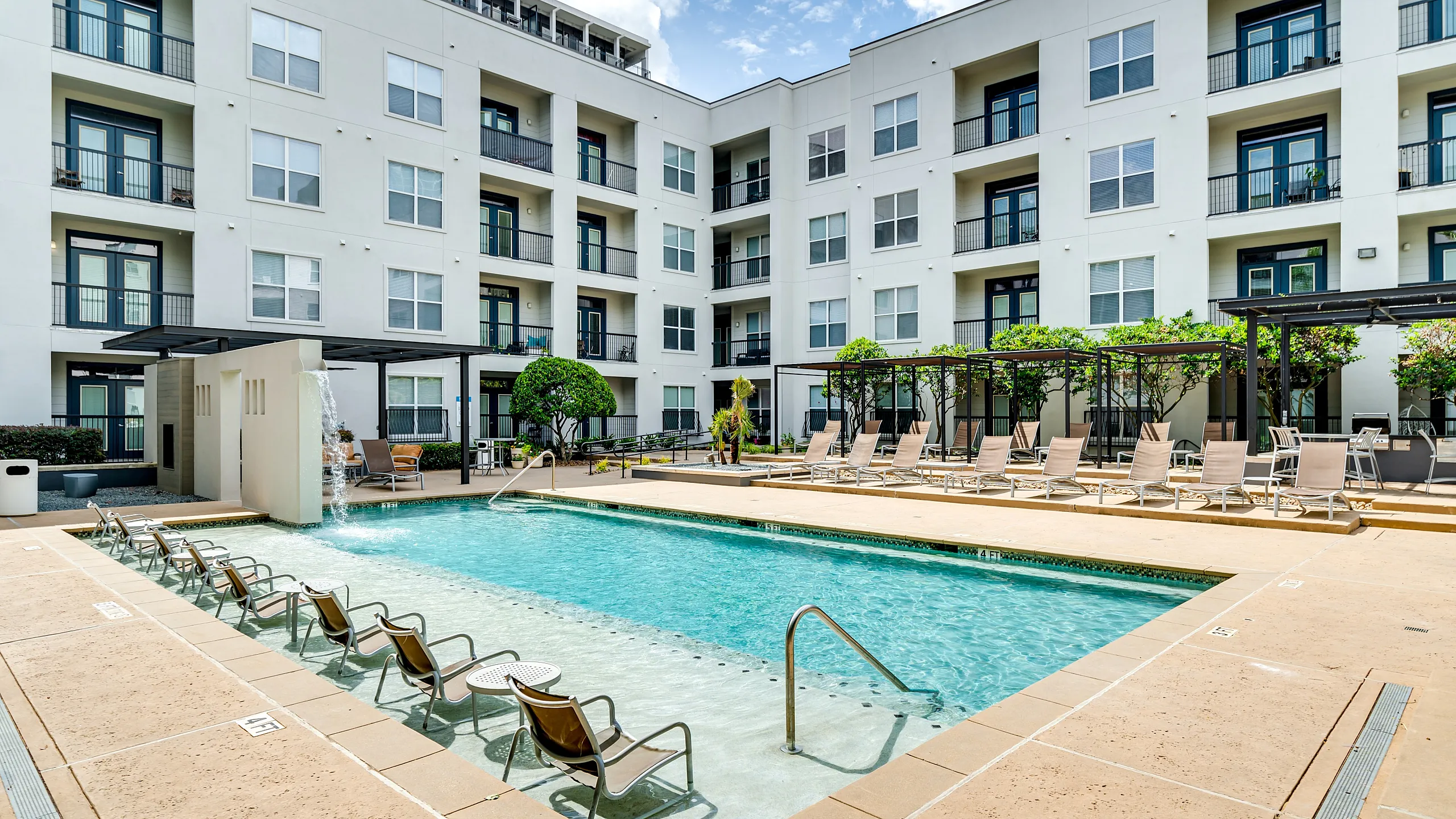 Contemporary apartment complex with a luxury pool area, featuring comfortable lounge chairs, cascading waterfall, and stylish landscaping for a perfect relaxation spot.