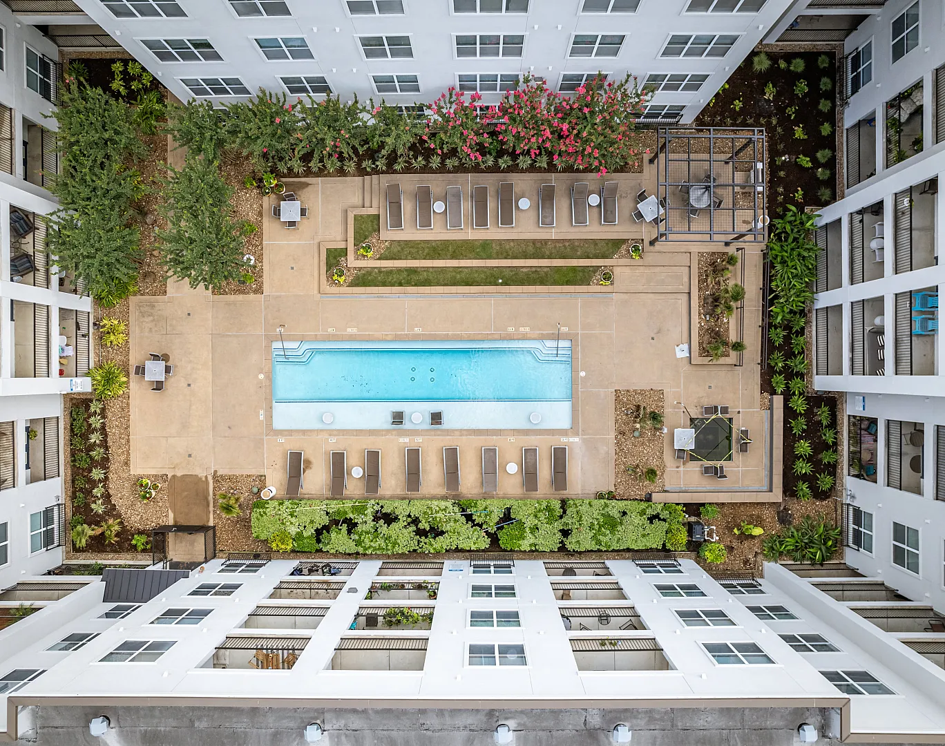 Bird's-eye view of a modern apartment courtyard with a pristine swimming pool, lush greenery, and comfortable lounge areas, framed by sleek white apartment buildings.