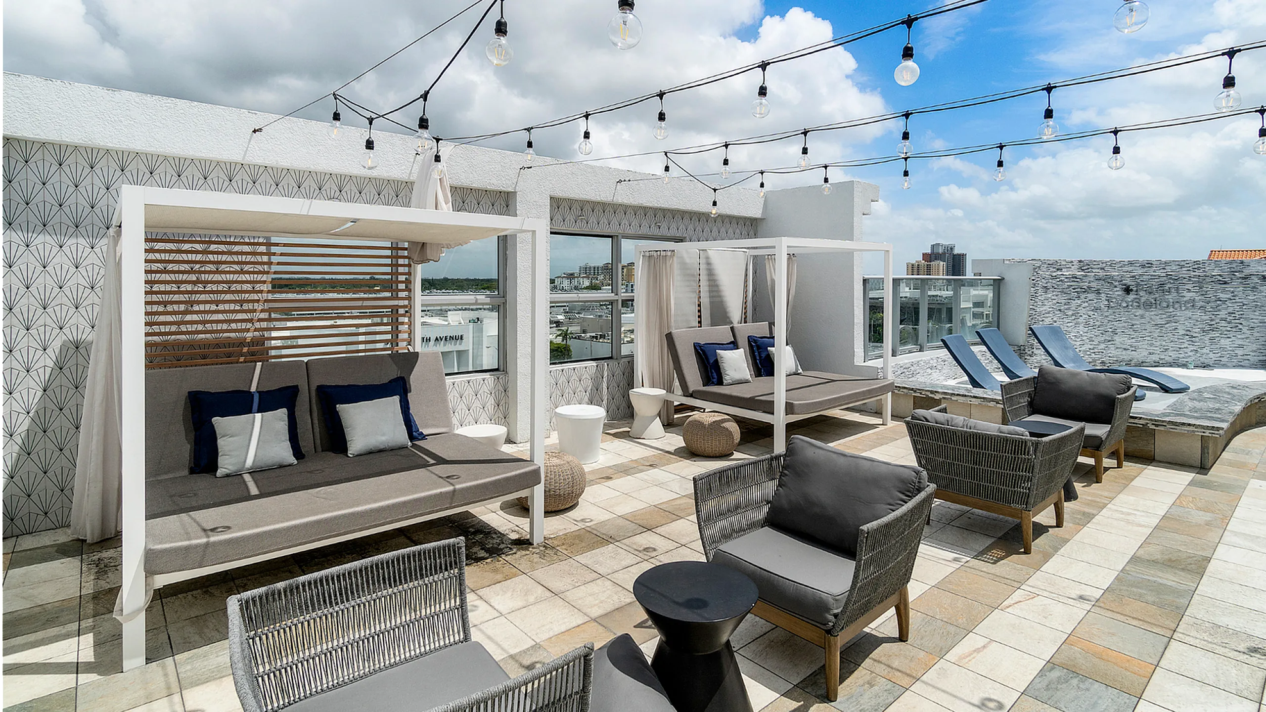 Chic rooftop terrace with cozy cabanas, stylish lounge chairs, and modern patio furniture under string lights against a vibrant blue sky.
