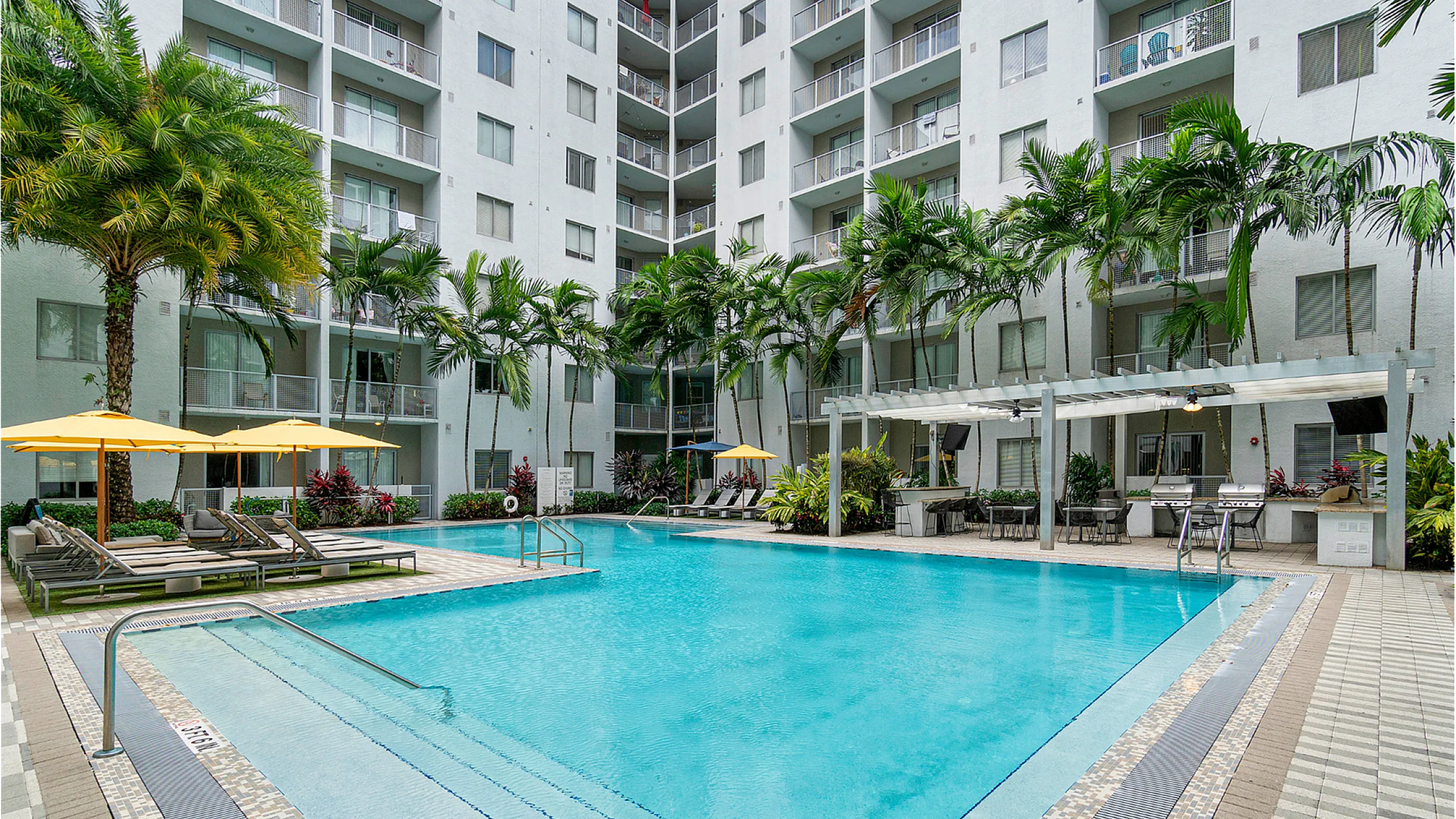 Luxurious modern apartment complex with a sparkling swimming pool, surrounded by lush palm trees and relaxing lounge chairs.