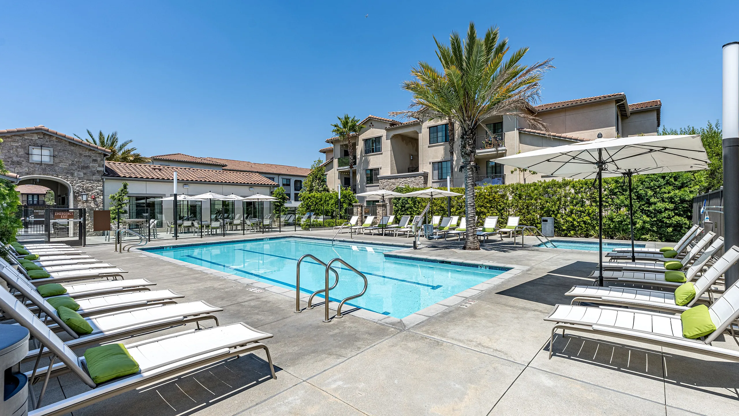 Beautiful apartment complex featuring a sparkling outdoor pool surrounded by lush greenery and modern lounge chairs, perfect for relaxation under a clear blue sky.