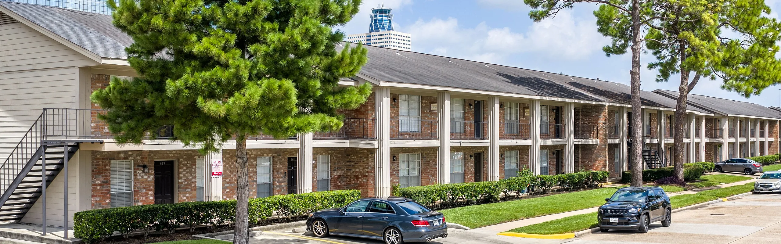 Charming apartment complex with a modern exterior, lush greenery, and ample parking spaces under a bright blue sky.