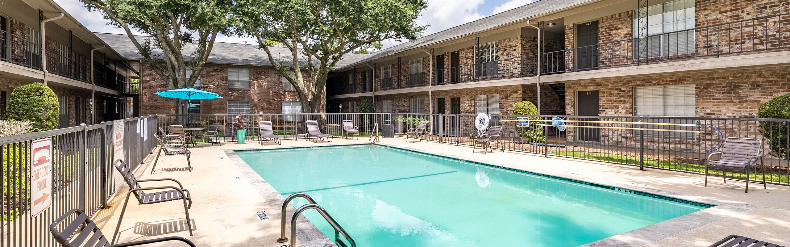 Inviting apartment pool area with sun loungers, surrounded by lush greenery and charming brick buildings, perfect for relaxation.