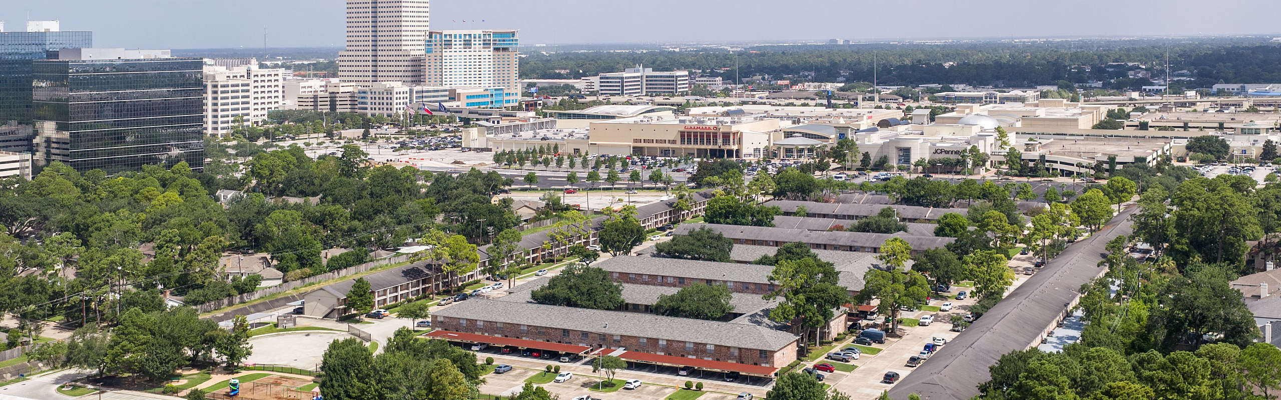 Vibrant aerial view of a lush suburban area with modern buildings and tree-lined streets, showcasing dynamic city life and scenic greenery under a clear blue sky.