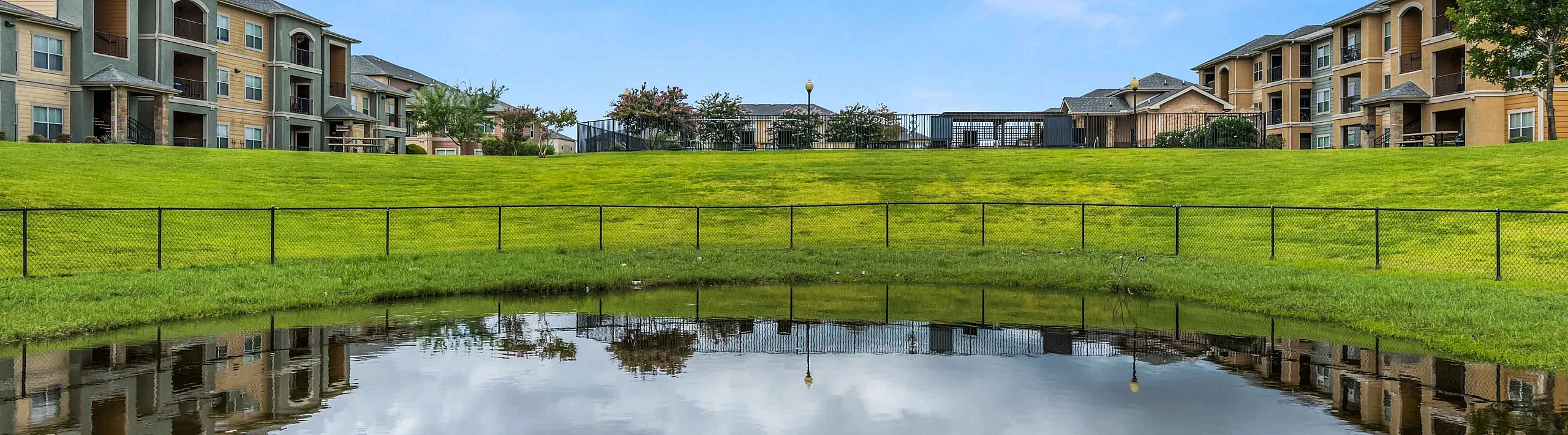 Pond reflecting the sky beside a fenced lawn with apartment buildings on each side
