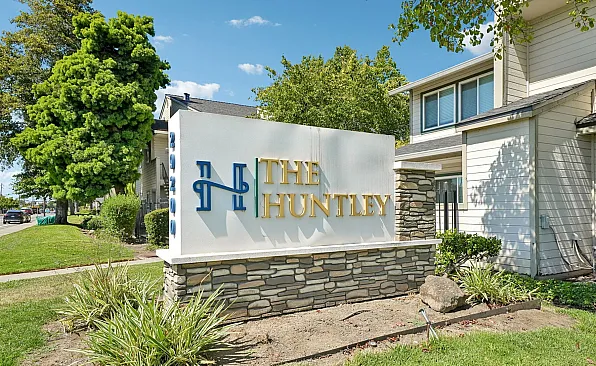 Inviting entrance to The Huntley Apartments with elegant signage and lush greenery, promising a serene living environment.