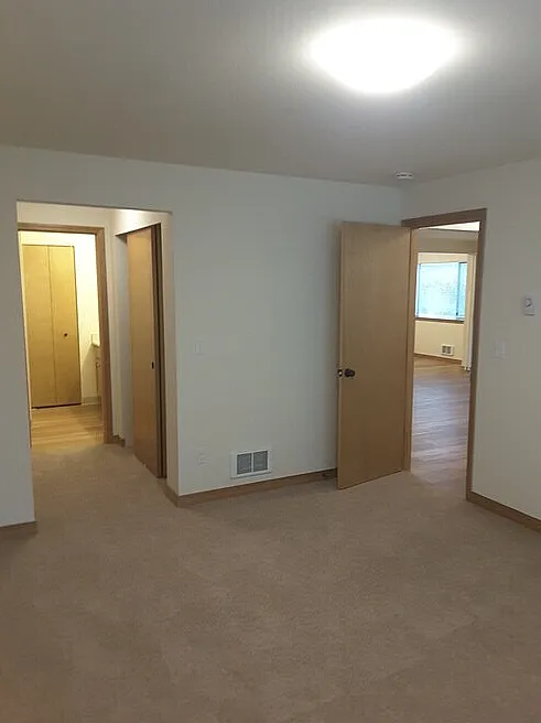 Bright and welcoming apartment interior featuring carpeted floors, wooden doors, and open doorways leading to spacious rooms with ample natural light.