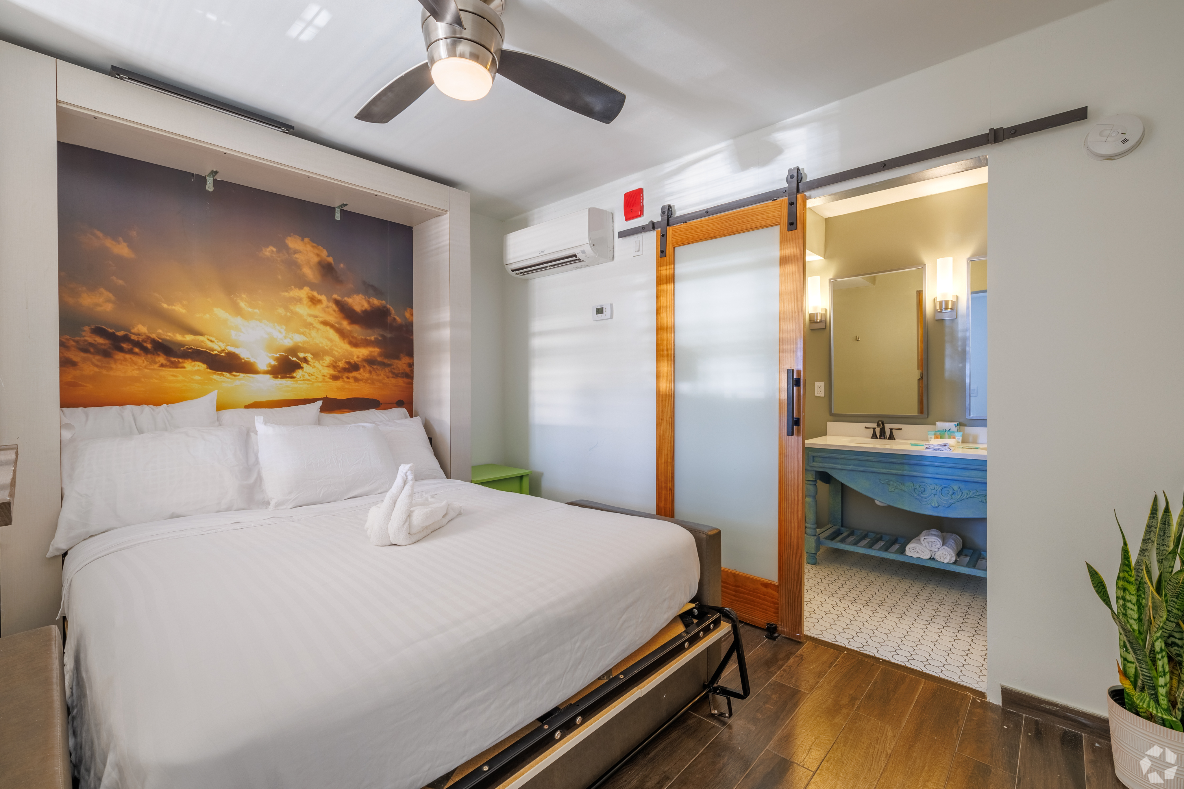 Cozy bedroom with elegant decor, featuring a plush bed, serene sunset mural, sleek ceiling fan, and a stylish sliding door leading to a chic bathroom.