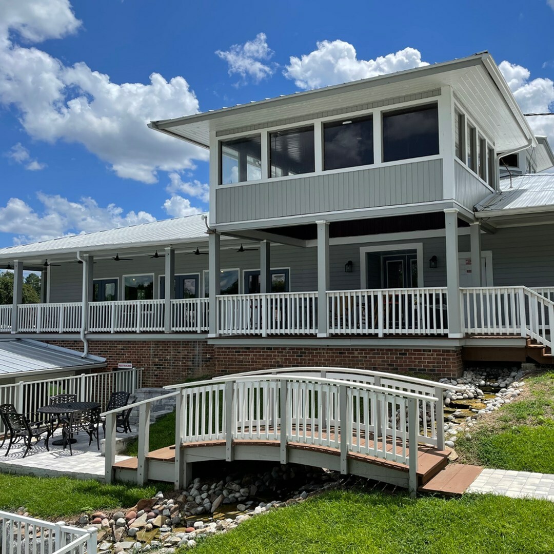 Captivating country-style home with expansive wraparound porch, charming wooden bridge, and elegant outdoor patio set amidst lush greenery.