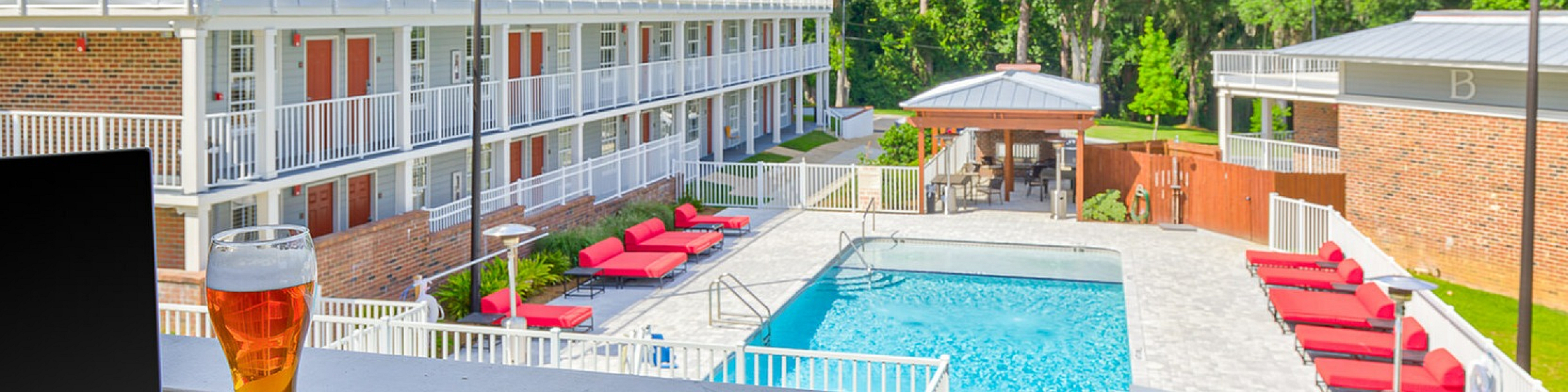 Charming apartment balcony view featuring a sparkling pool, red sun loungers, and lush greenery under a bright blue sky.