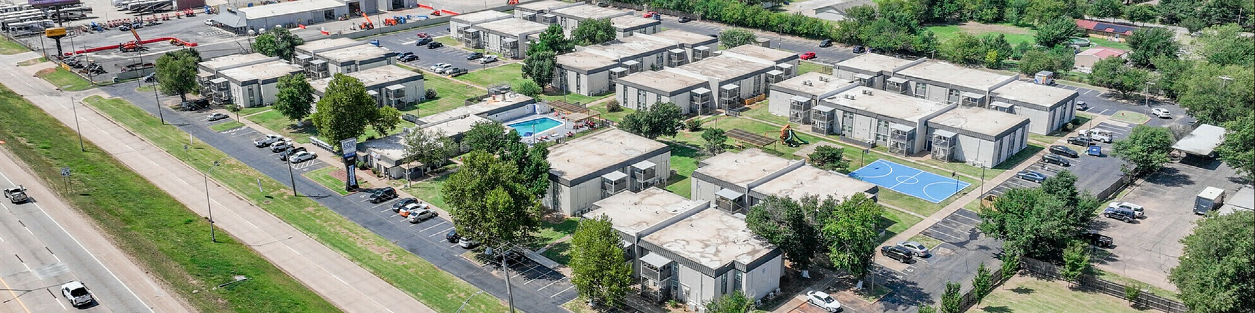 Scenic aerial view of a modern apartment complex featuring lush greenery, a sparkling swimming pool, and a basketball court, all conveniently located near a major roadway.