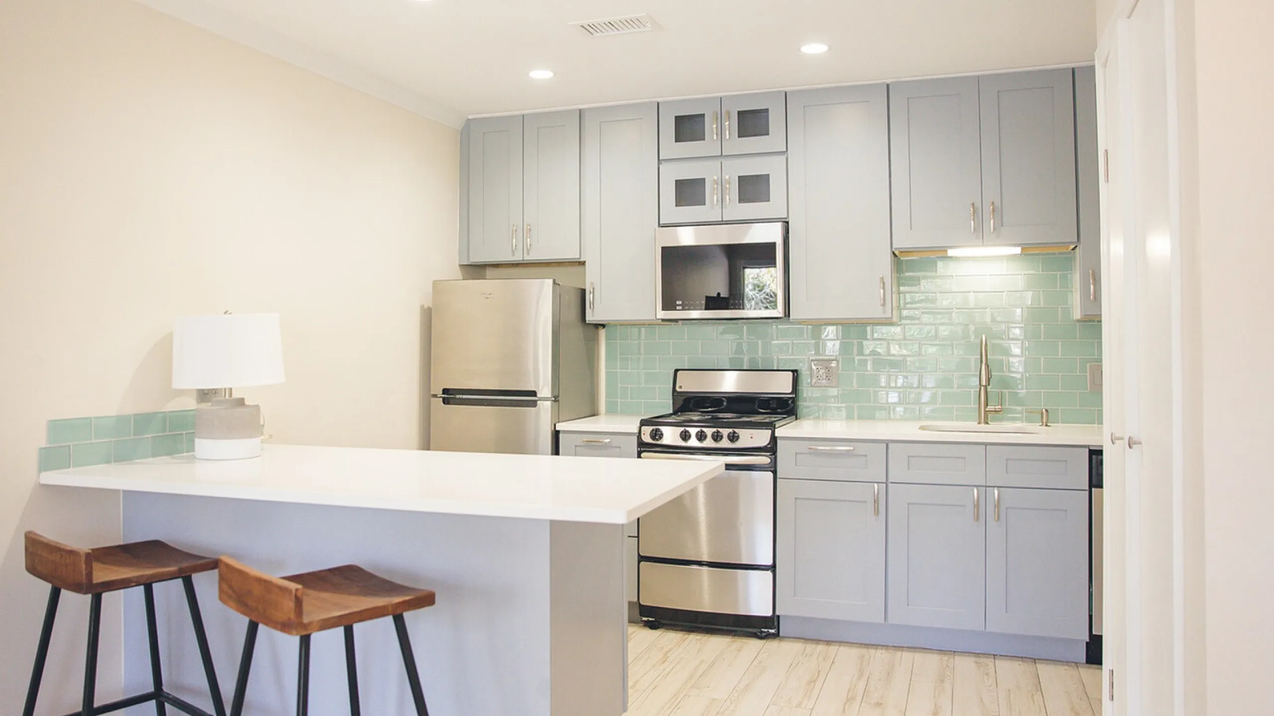 Modern kitchen with sleek stainless steel appliances, soft gray cabinetry, and a charming breakfast bar with two stools, featuring chic mint-green backsplash tiles.