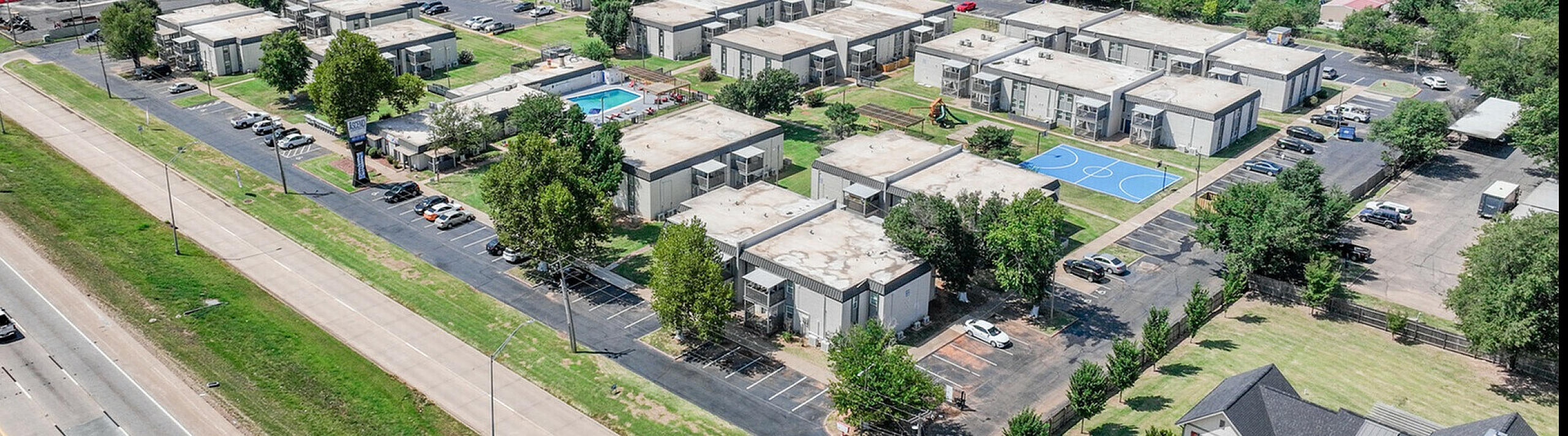 Expansive aerial view of a vibrant apartment community featuring multiple buildings with green spaces, a sparkling swimming pool, and a basketball court, set against a backdrop of lush trees and blue skies.