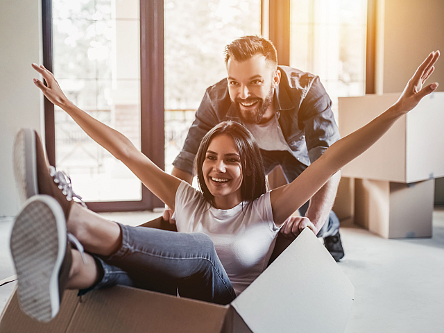 Joyful couple celebrating their new home by playing with moving boxes in a sunlit apartment.